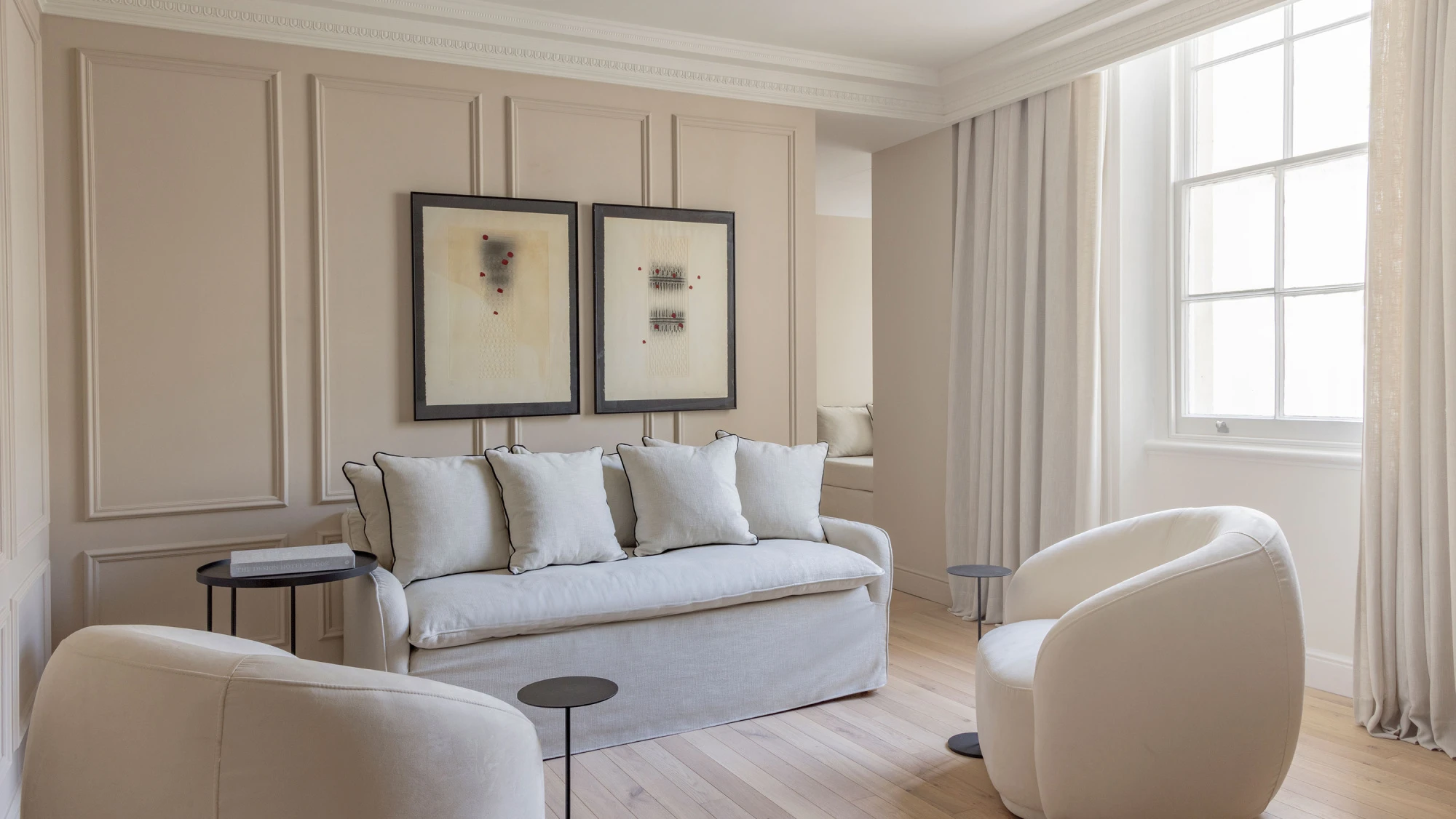 Sala de estar con sofá claro, sillones blancos y cuadros en la pared junto a una ventana con cortinas.
