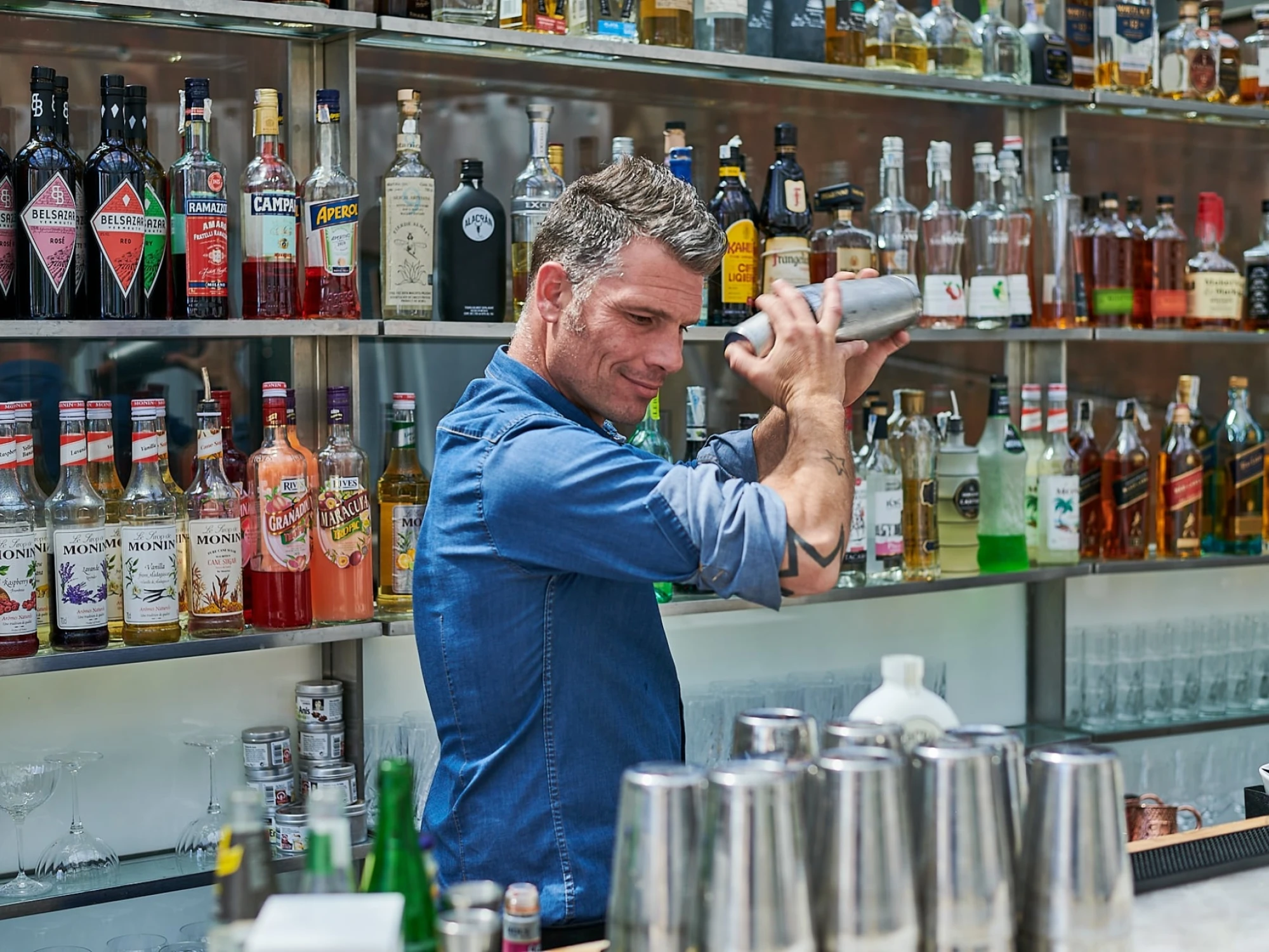 Un camarero sirve bebidas detrás de una barra con botellas y vasos en estanterías.