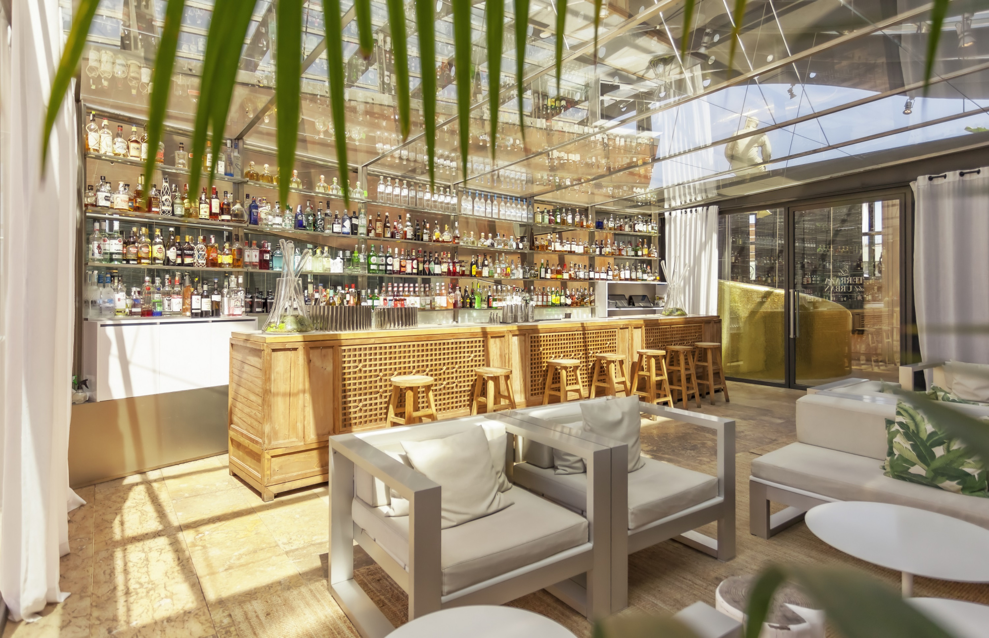 Se ve una barra con botellas alineadas y taburetes, junto a mesas y sillas en un espacio tipo bar con luz natural.