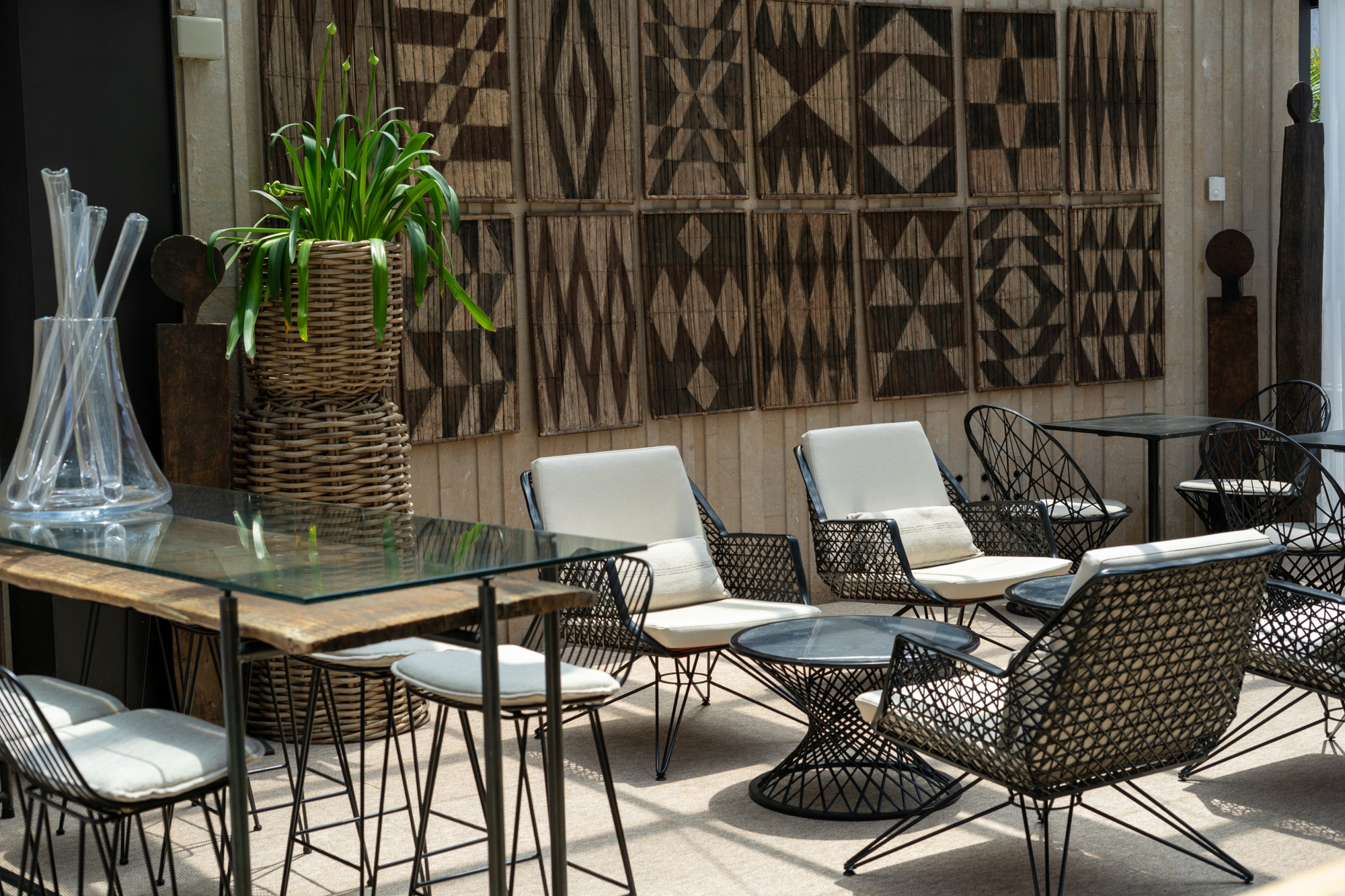 Terraza exterior con mesas de madera, sillas de metal y una planta en maceta junto a un panel decorativo.