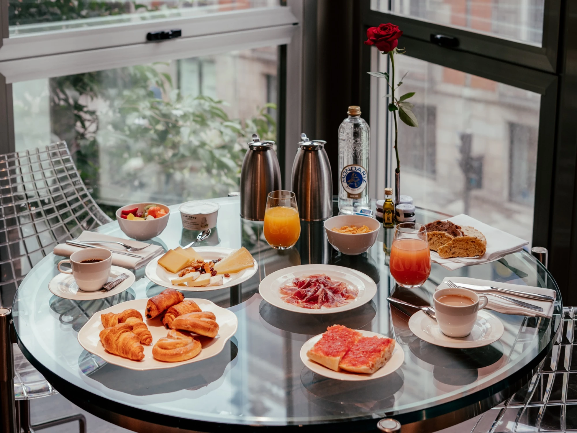 Se ve una mesa con platos de desayuno, fruta y bebidas sobre una bandeja junto a una ventana.