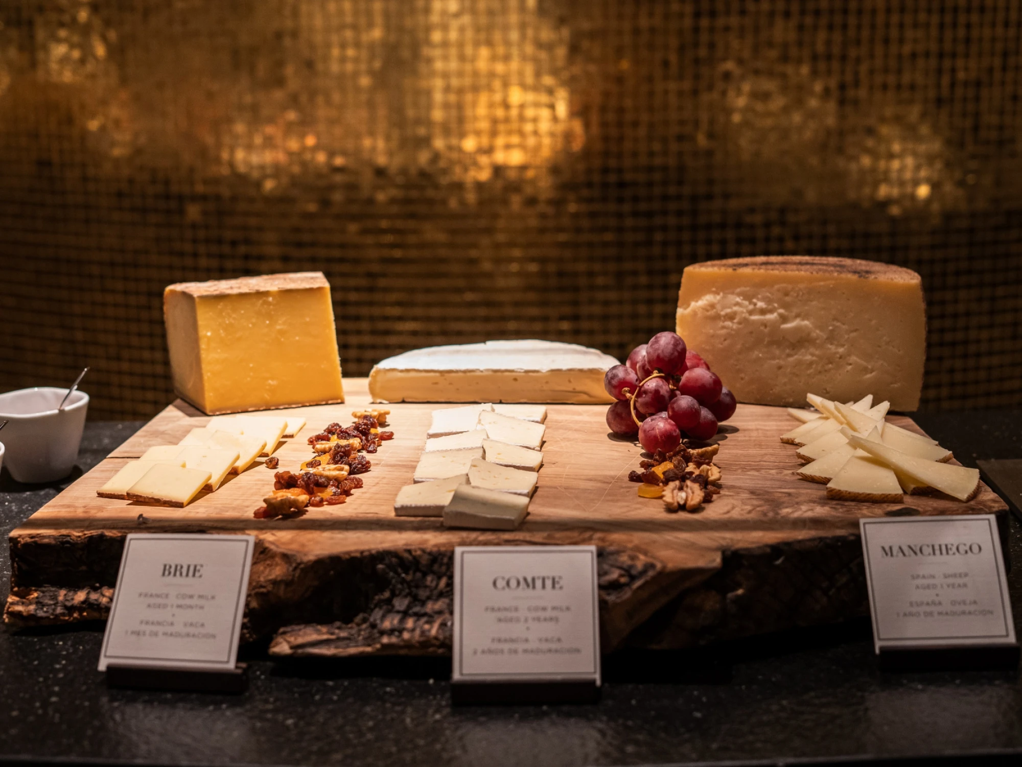 Se ve una mesa con tablas de queso, embutidos y uvas sobre un fondo de pared con iluminación cálida.