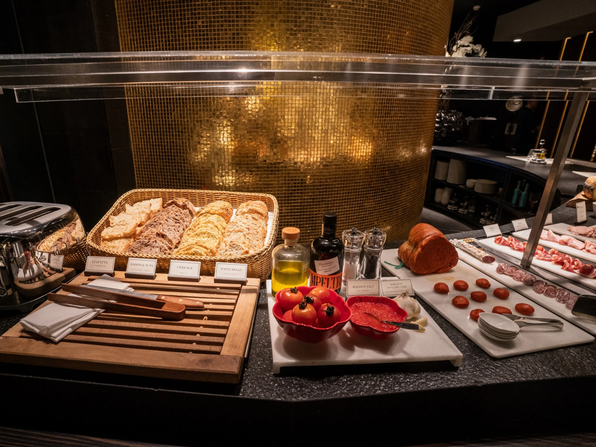 Se ve un buffet con bandejas de pan, frutas y recipientes sobre una mesa con tapa de cristal.