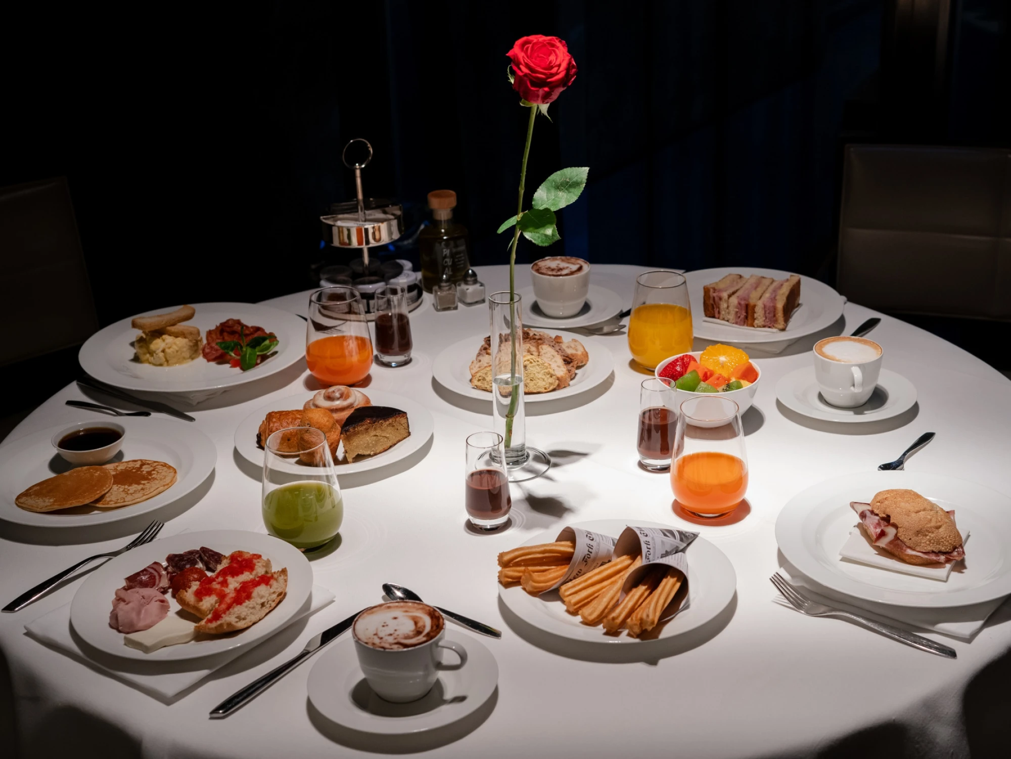 Se ve una mesa con platos variados, bebidas y una rosa en un jarrón sobre un mantel blanco.