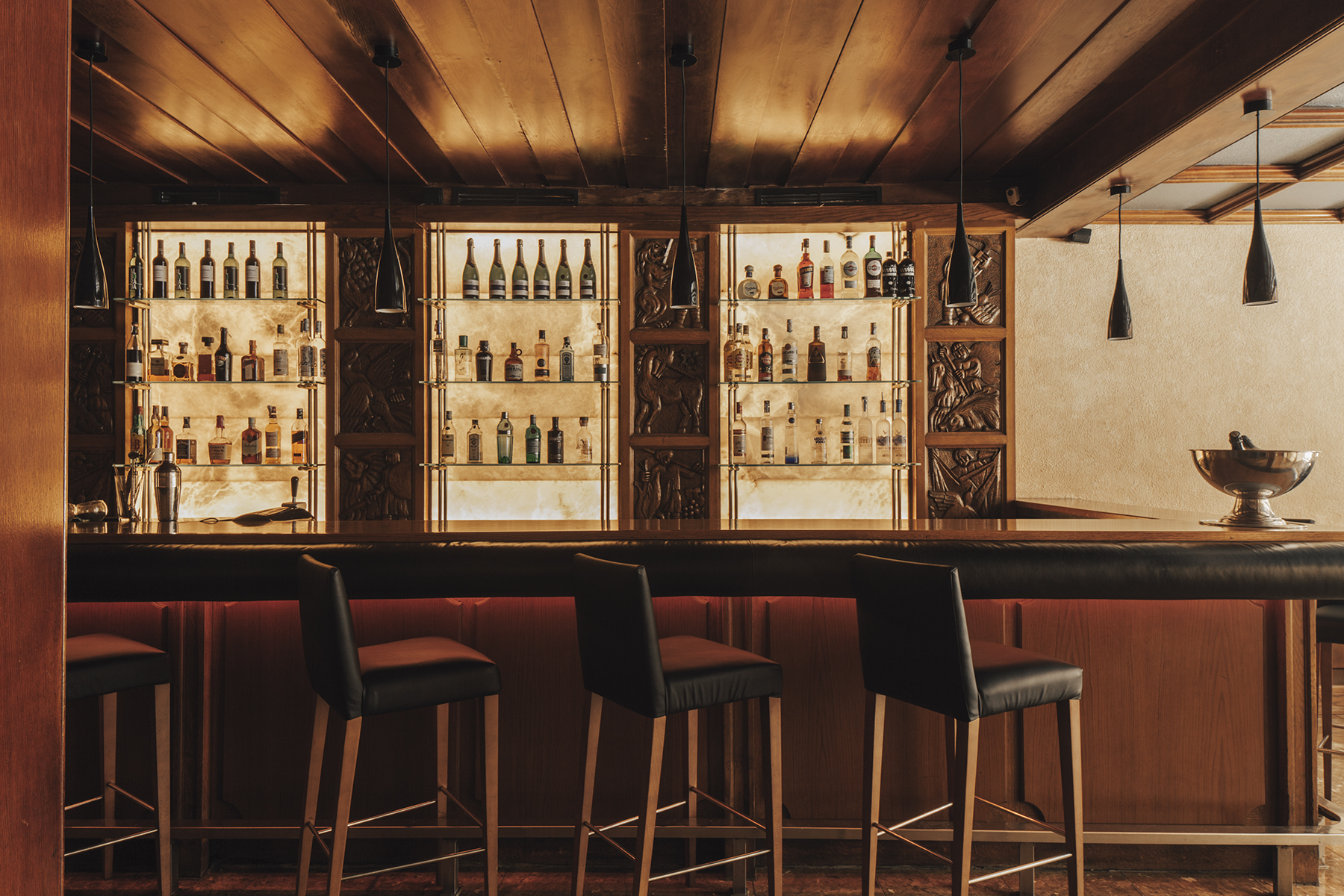 Se ve una barra con taburetes y botelleros iluminados en un bar interior.