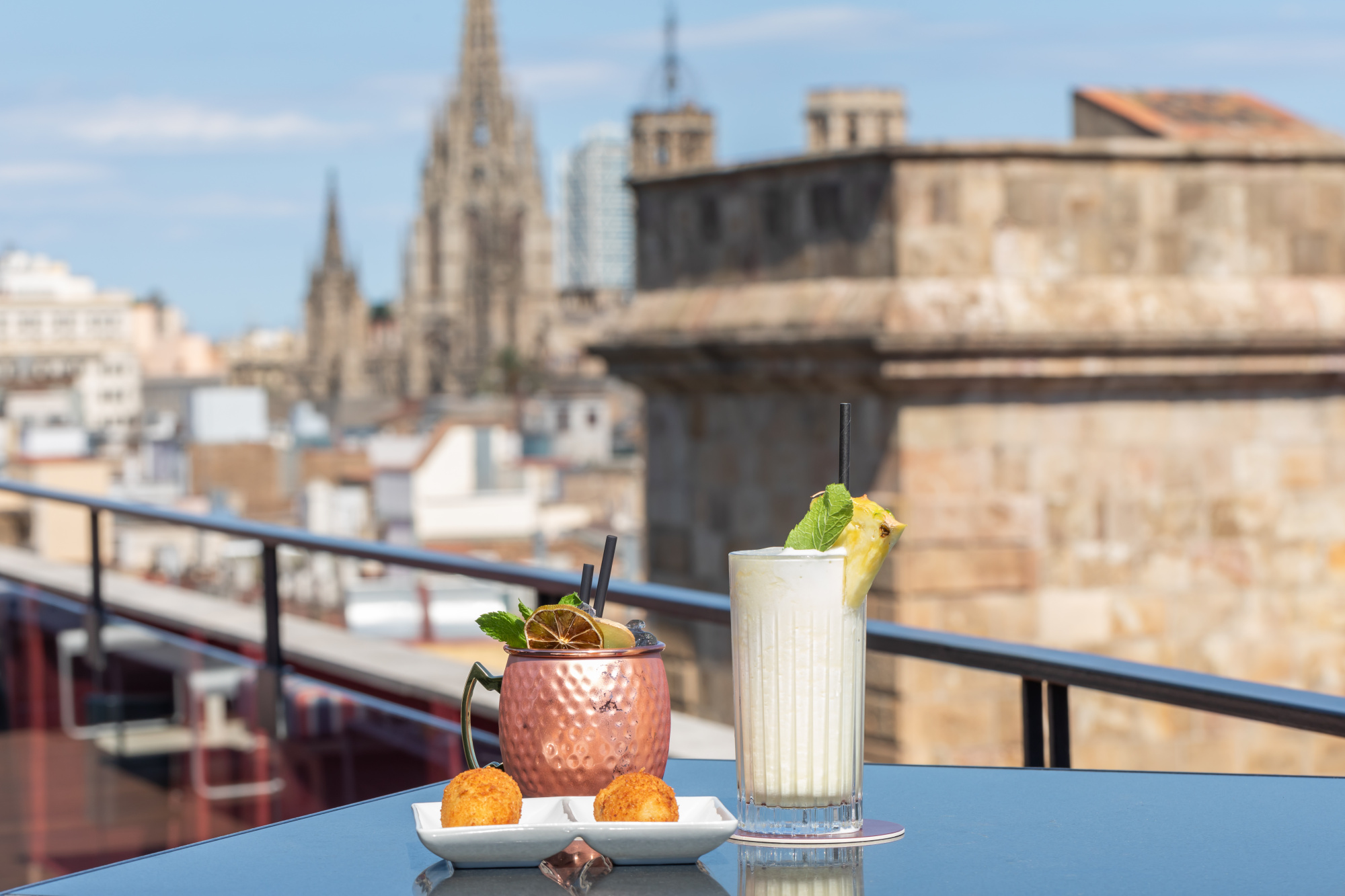 Auf einer Terrasse sieht man einen Tisch mit einem Tablett voller Früchte und Gläsern auf einem Geländer, im Hintergrund sind Gebäude zu sehen.