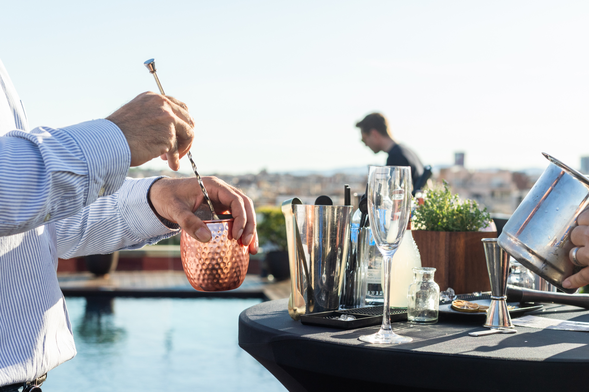 Se ven unas manos preparando bebidas con una coctelera y vasos sobre una mesa en una terraza al aire libre.
