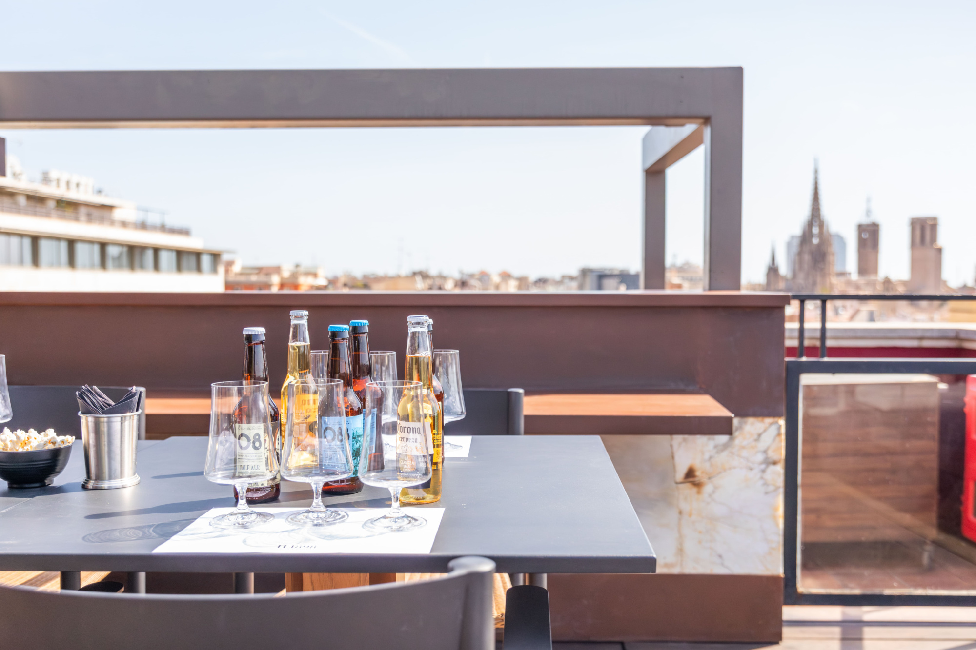 Se ve una mesa con botellas y vasos sobre una terraza, con barandilla y chimenea al fondo.
