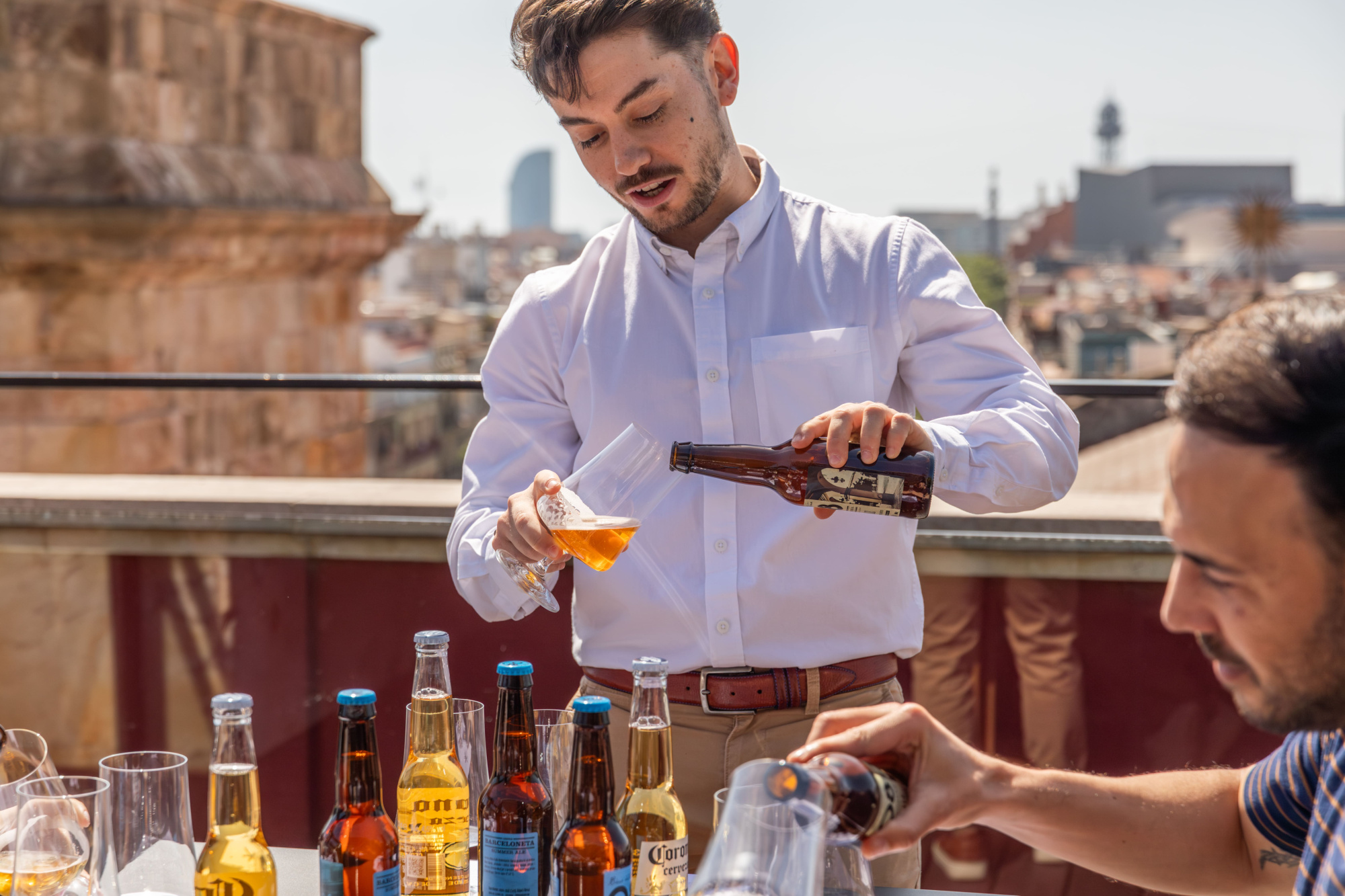 Un hombre sirve bebidas desde una mesa con botellas y vasos en una terraza al aire libre.