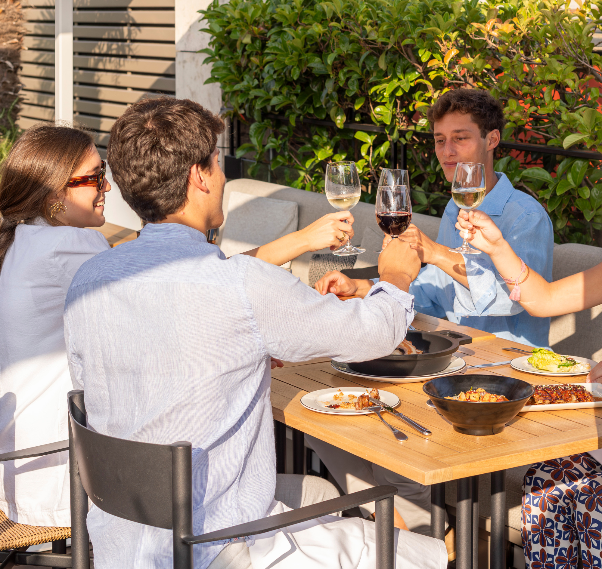 Cuatro personas brindan con copas alrededor de una mesa con comida en una terraza al aire libre.
