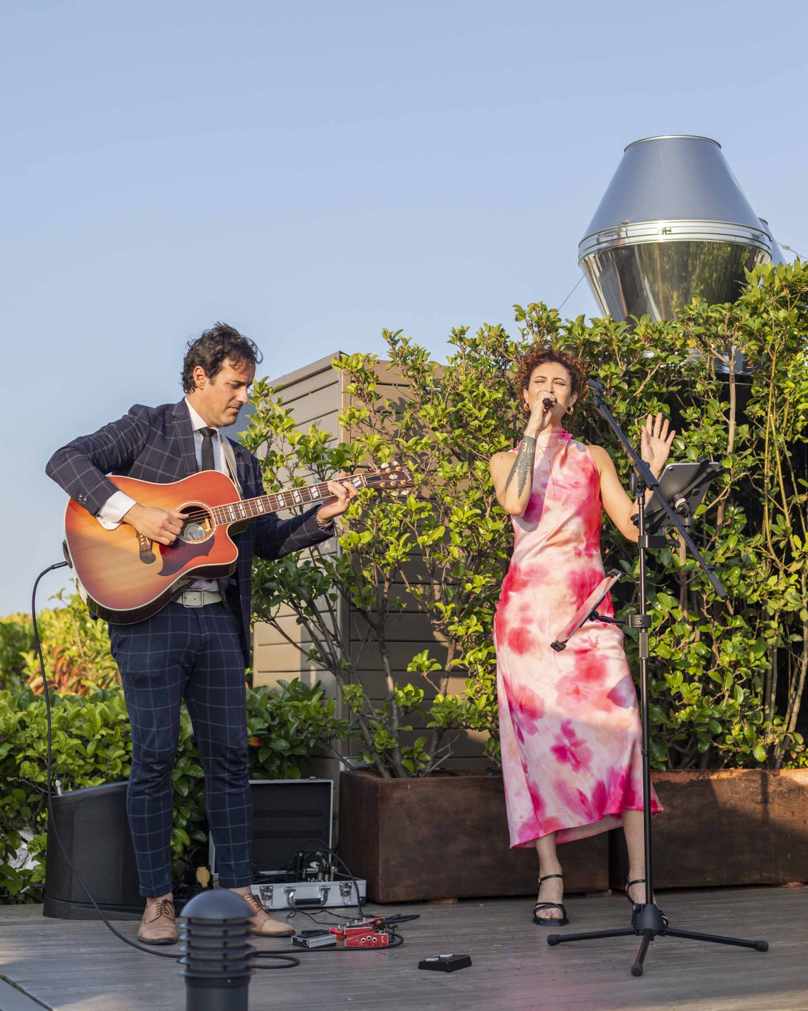 On an outdoor stage, two musicians perform with a singer and an acoustic guitar.
