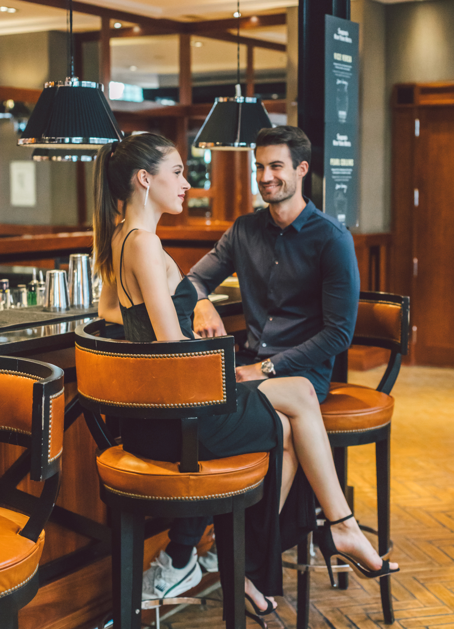 Una pareja conversa sentada en sillas de un bar con barra y taburetes al fondo.