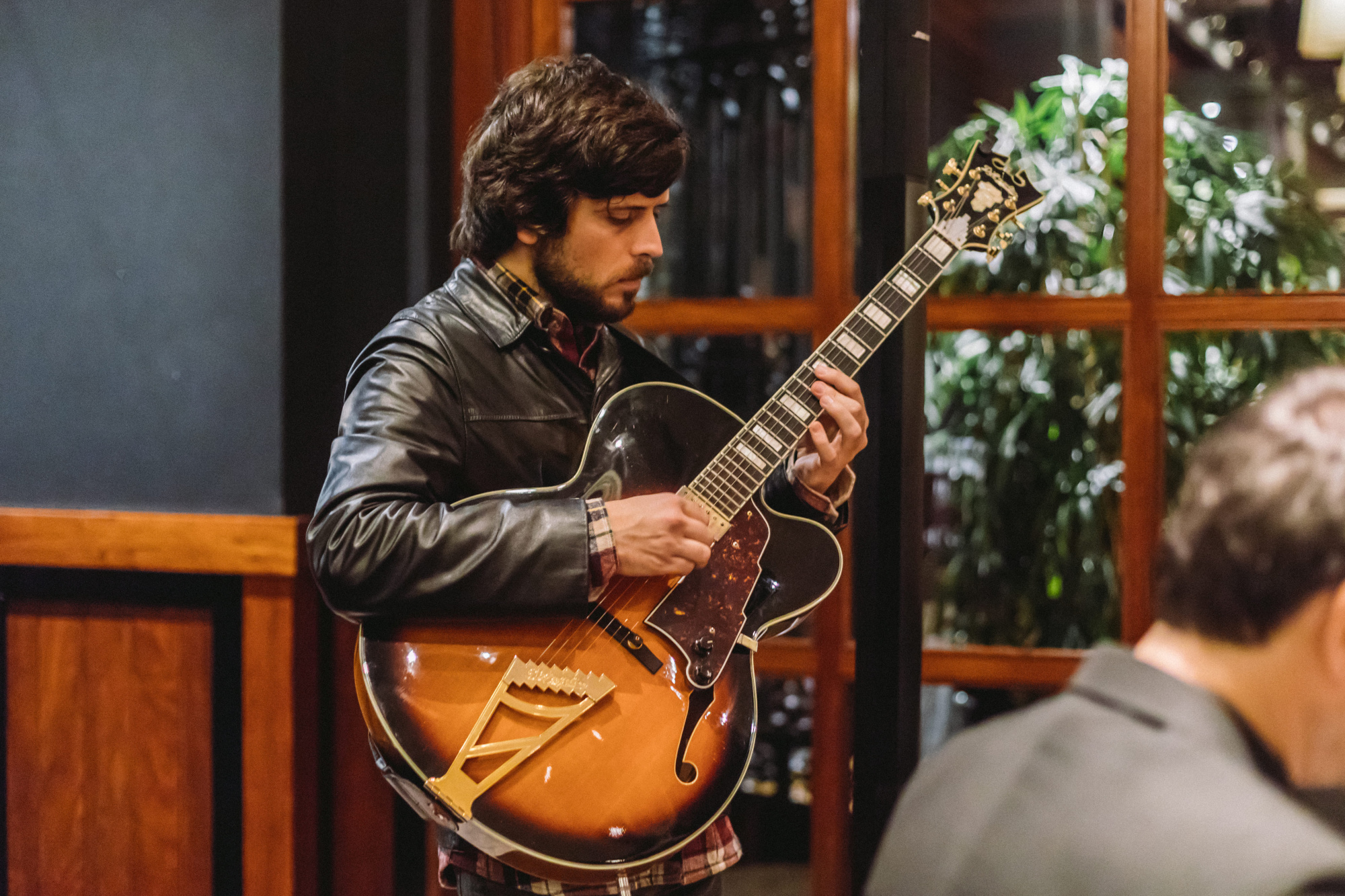 Un hombre toca una guitarra acústica en un espacio interior con otras personas sentadas cerca.