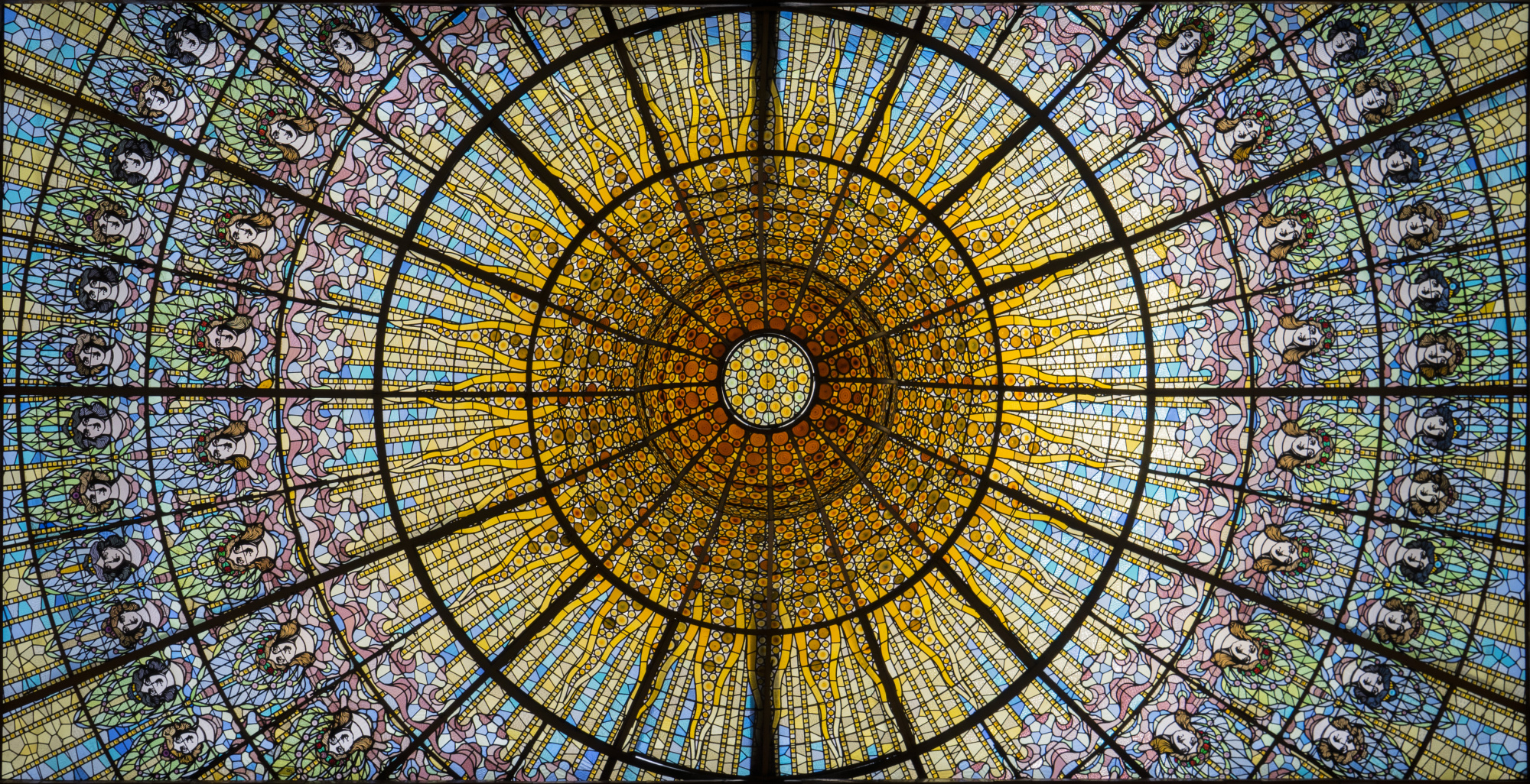 Describe una vidriera modernista circular con motivos florales y un marco negro en el techo.