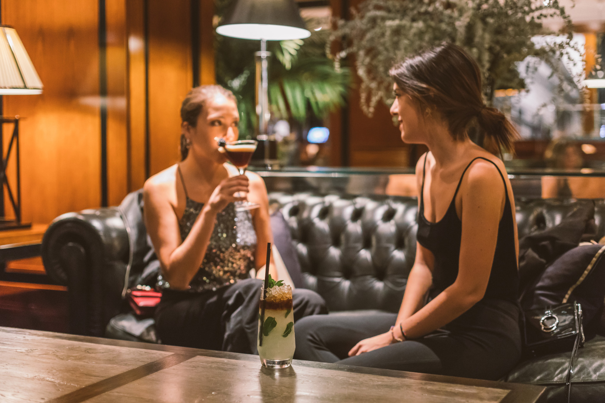 Dos personas conversan sentadas en un bar con bebidas y un jarrón sobre la mesa.