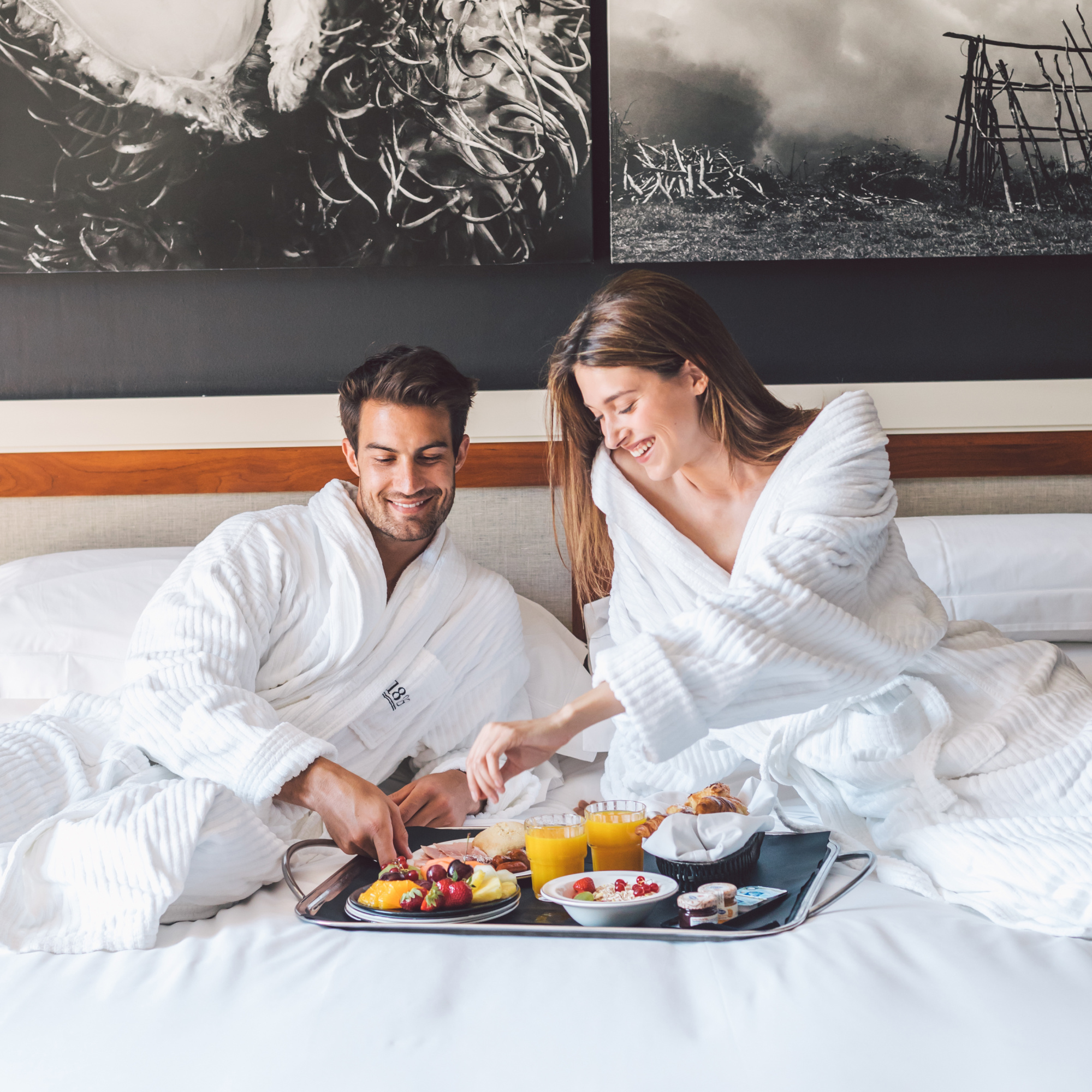 Una pareja desayuna en una bandeja sobre una cama con sábanas blancas.