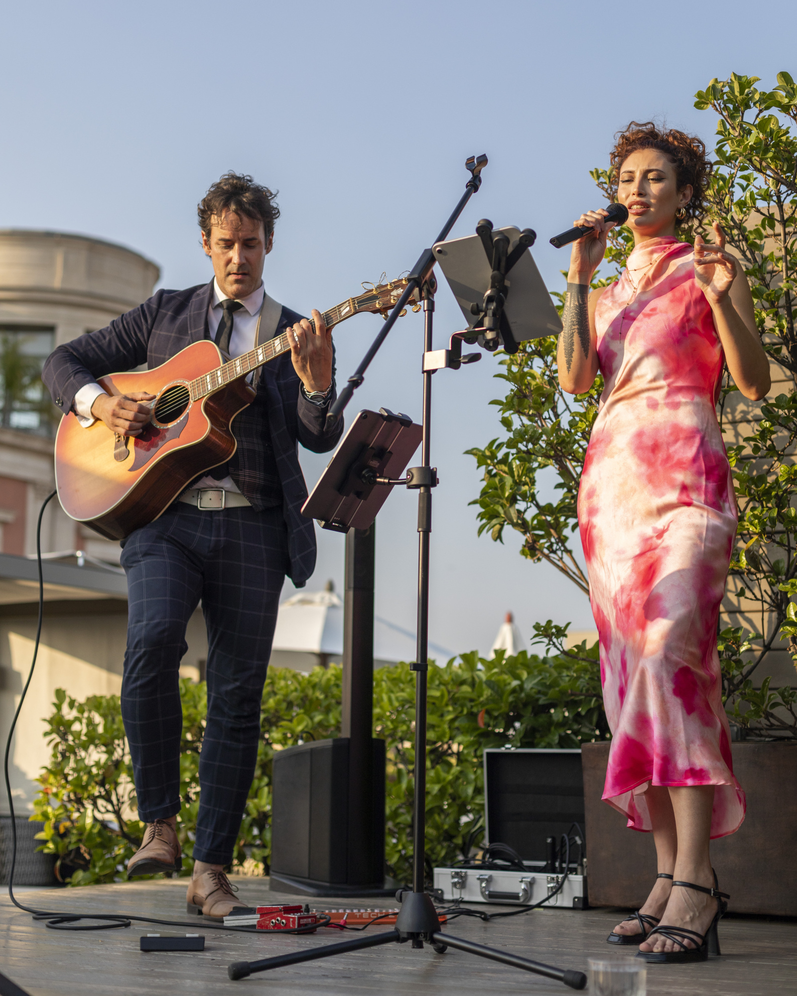 Una cantante y un guitarrista actúan en un escenario al aire libre con micrófono y equipo de sonido.