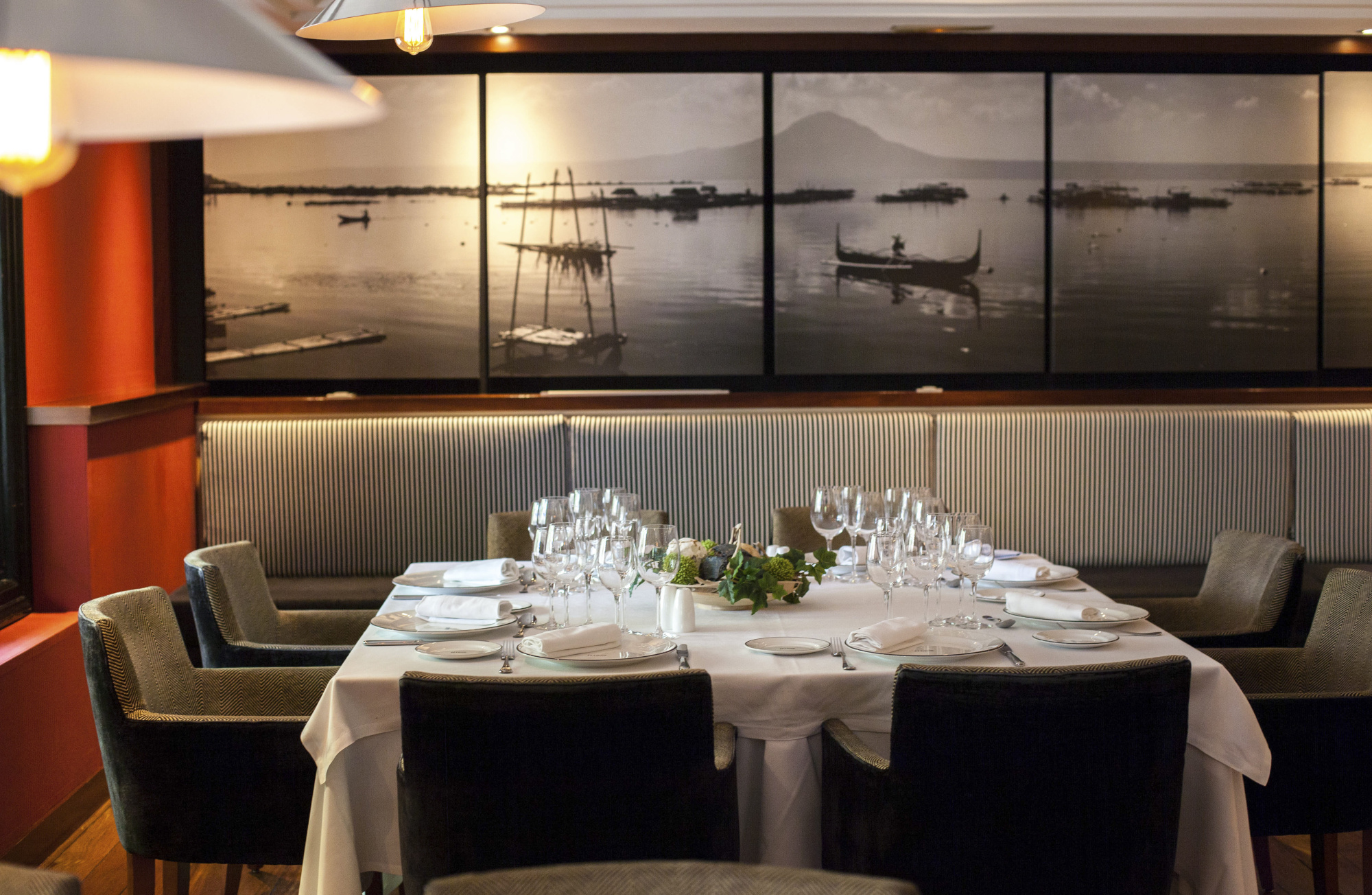 Una mesa puesta con copas y platos se ve en un comedor con asientos tapizados y decoración marina en la pared.