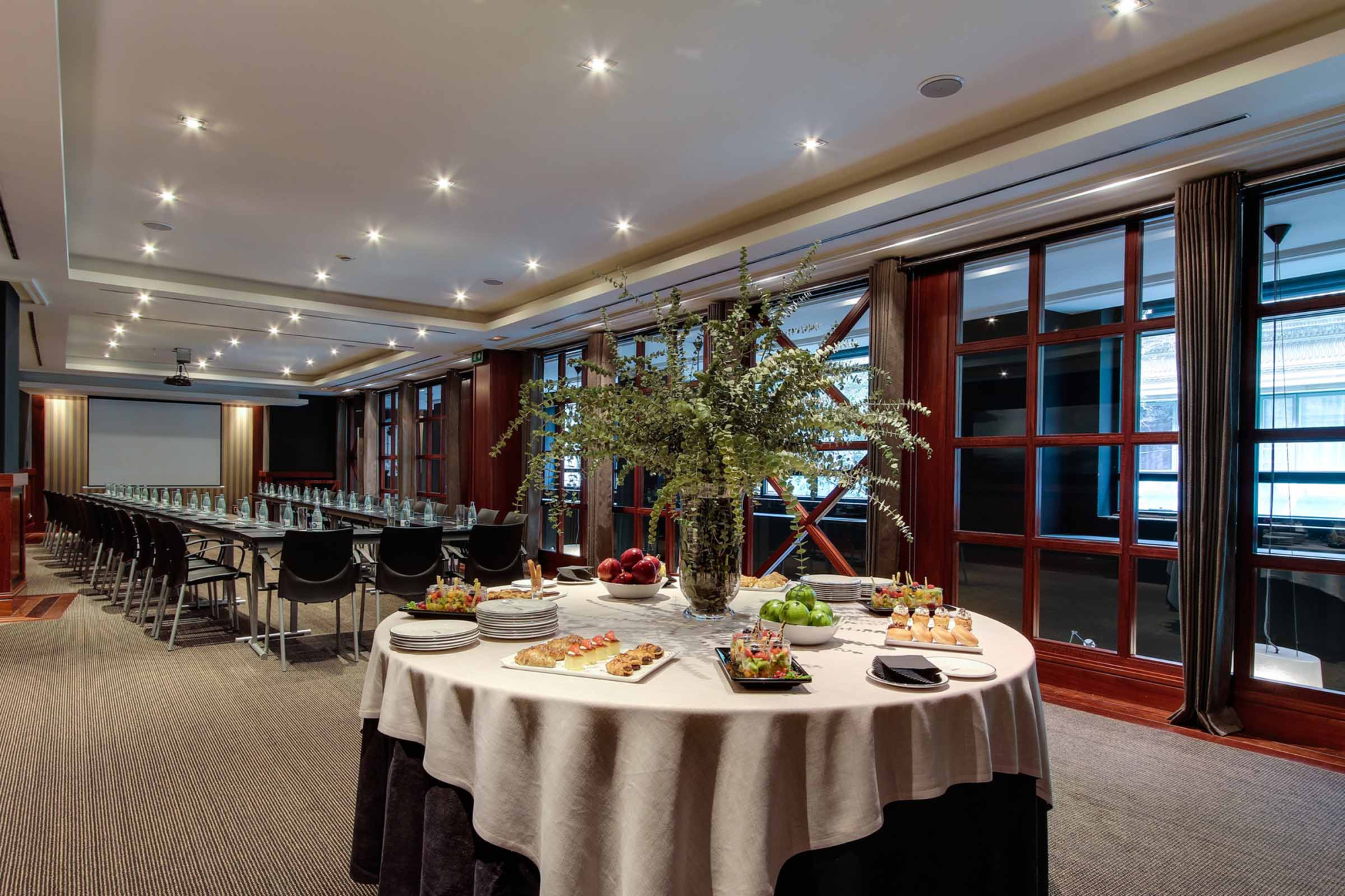 Se ve un comedor con mesas cubiertas, sillas y ventanales grandes con luz interior.