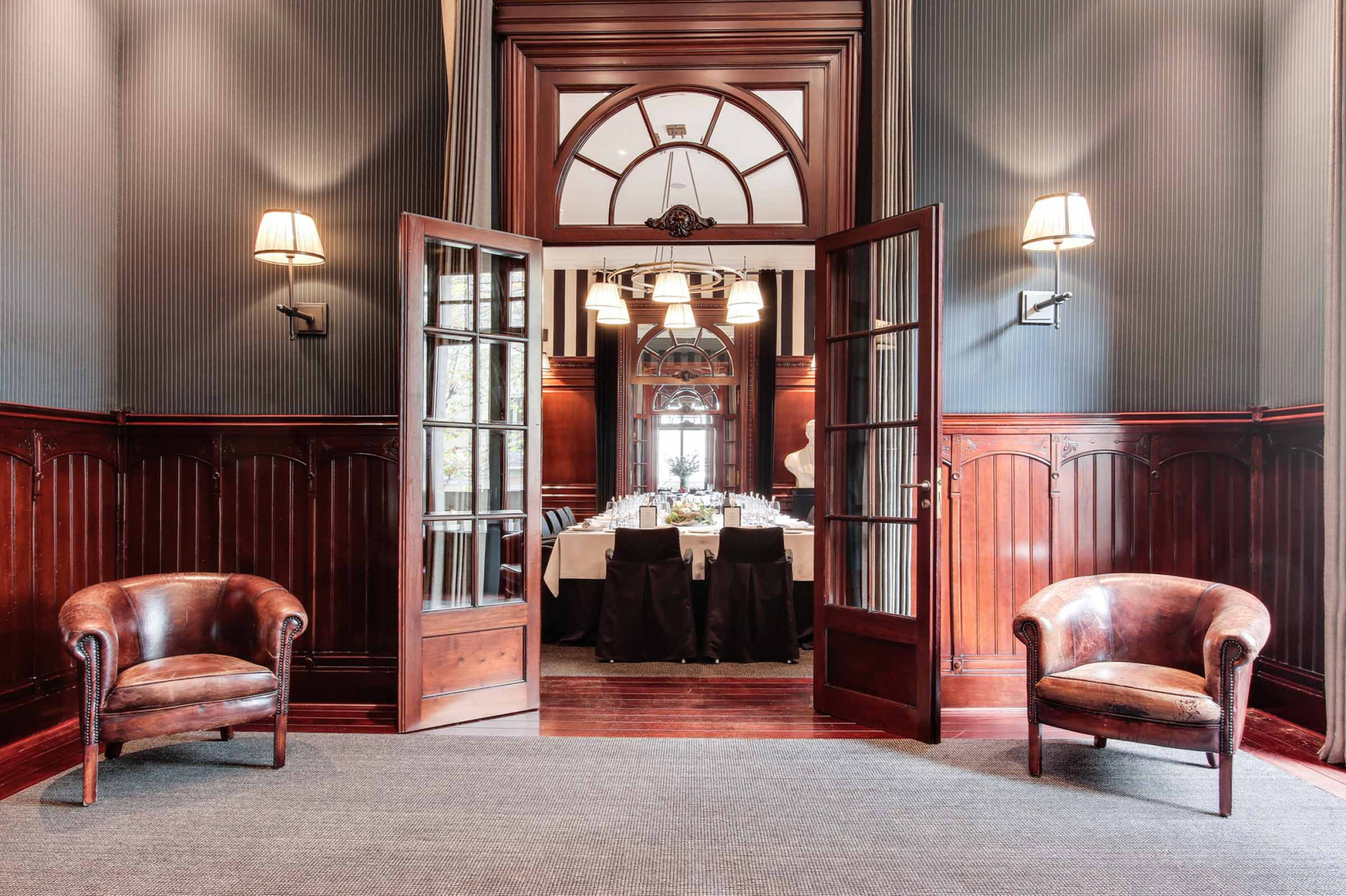 La imagen muestra una sala con dos sillones de cuero, lámparas de pared y una puerta abierta hacia un comedor.