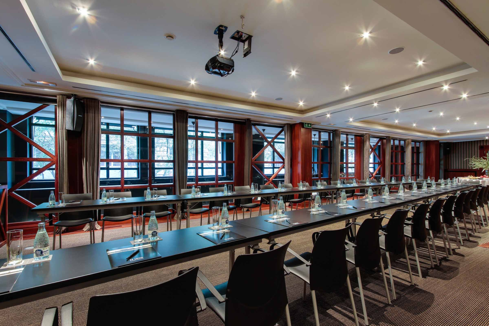 La imagen muestra una sala de reuniones con mesas largas, sillas y grandes ventanales.
