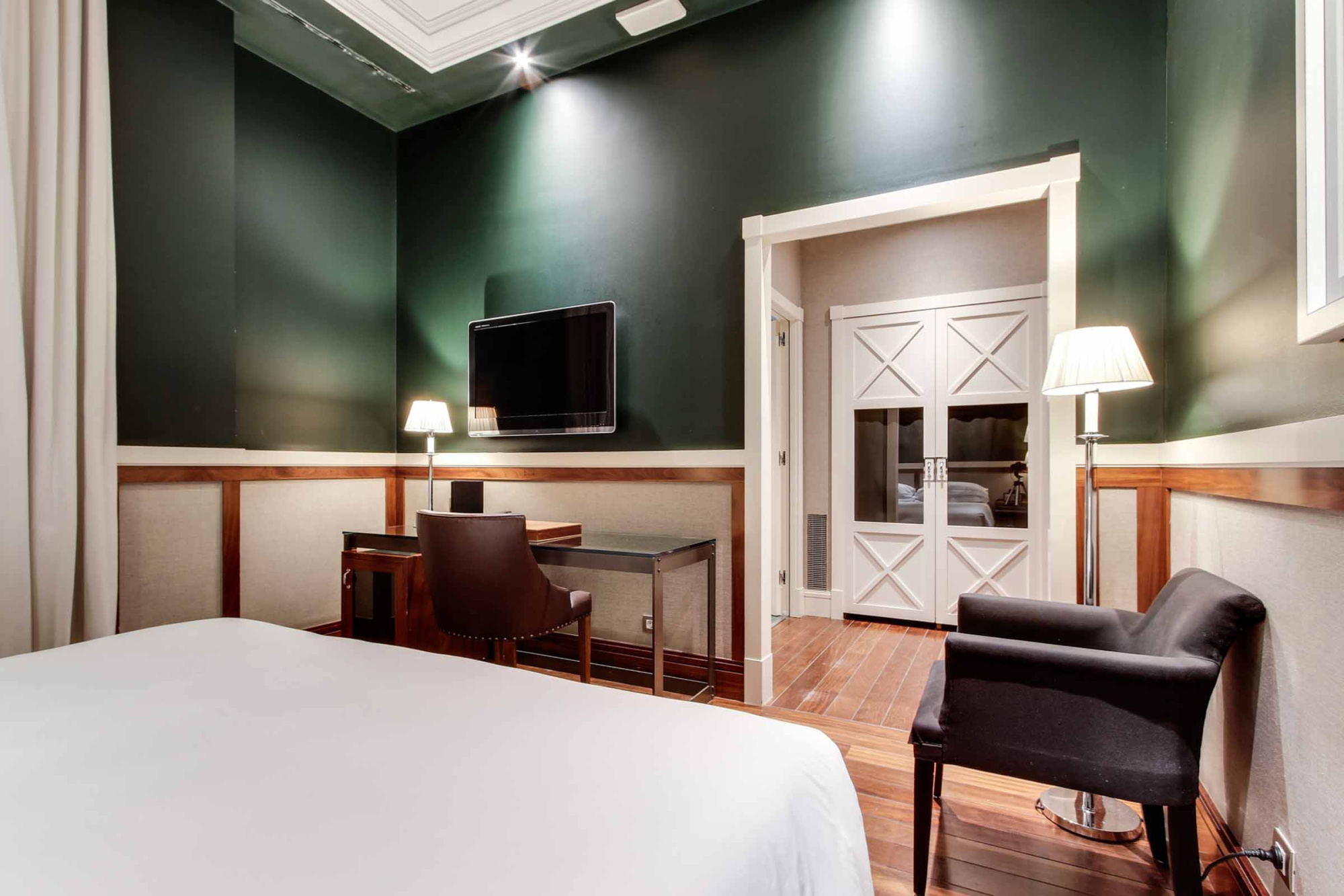 Se ve una cama, un escritorio con silla y una televisión en una habitación con paredes verdes y lámparas de pie.
