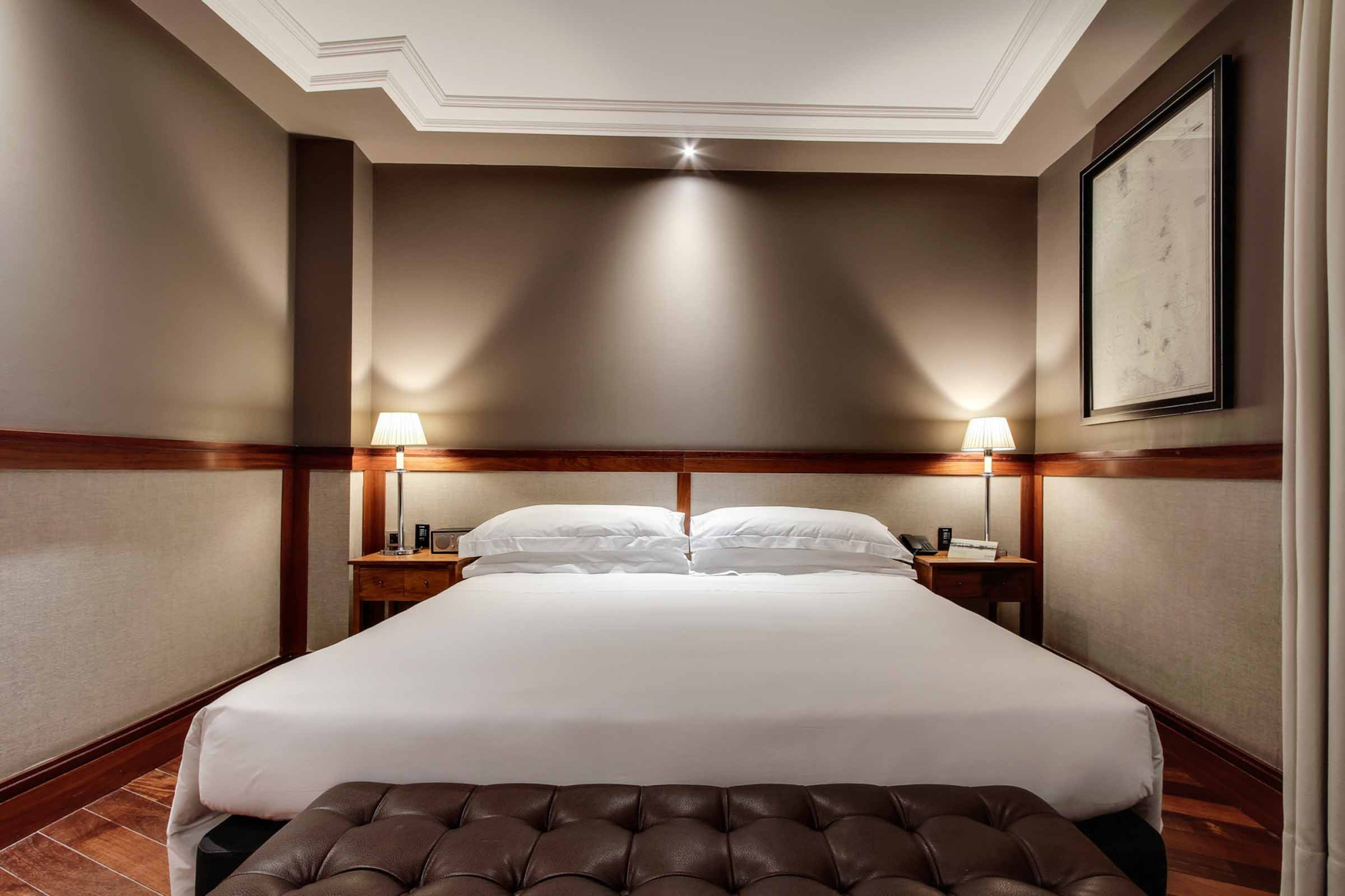 Se ve una cama doble con cabecero acolchado y dos mesitas de noche en una habitación con paredes beige.