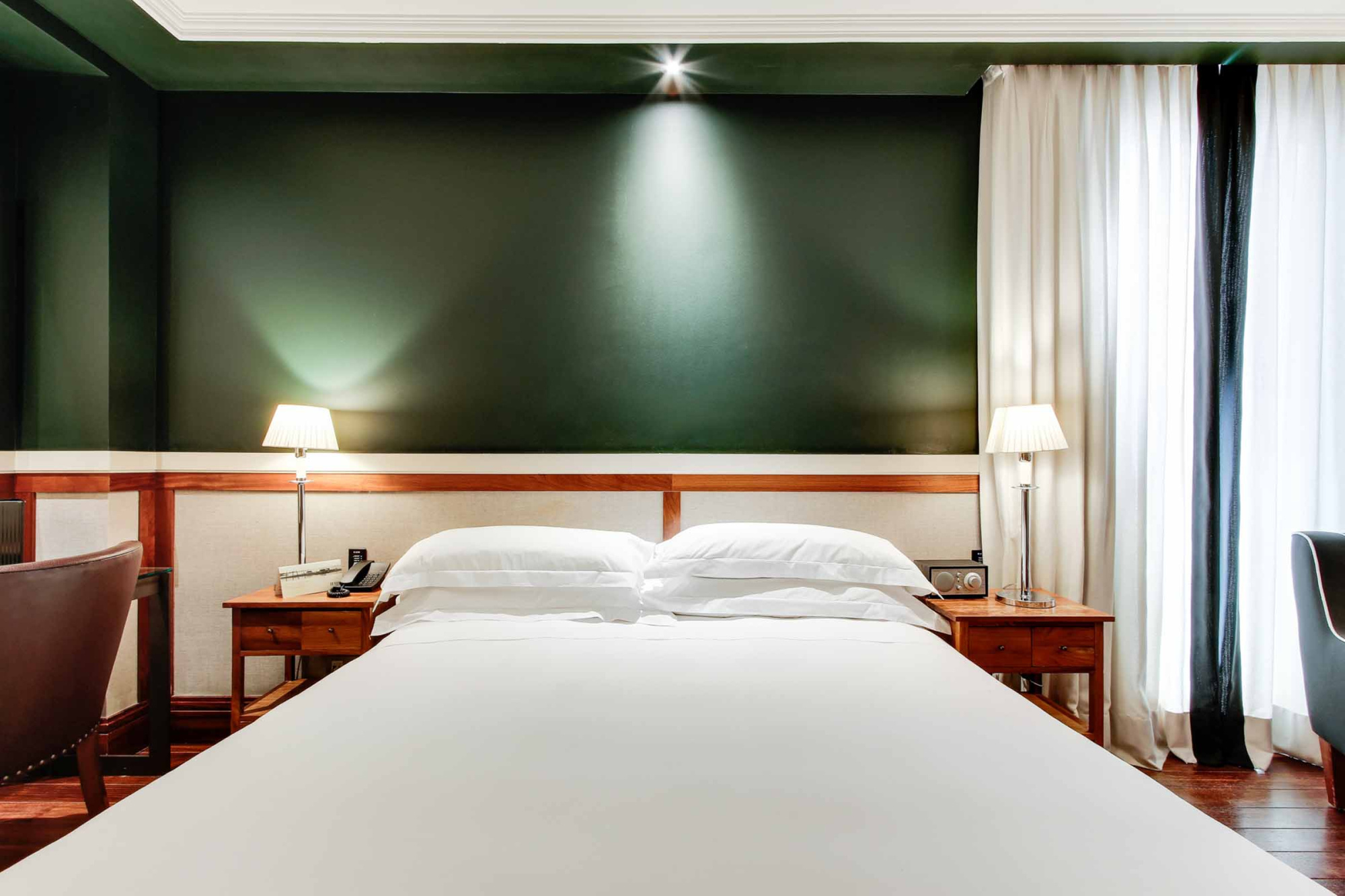 Si vede un letto matrimoniale con comodini e lampade, insieme a tende e una parete verde nella stanza.
