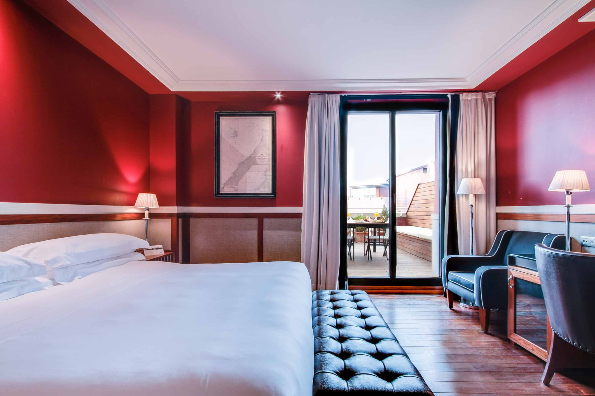 La imagen muestra una habitación con cama, banco tapizado y una puerta hacia el balcón.