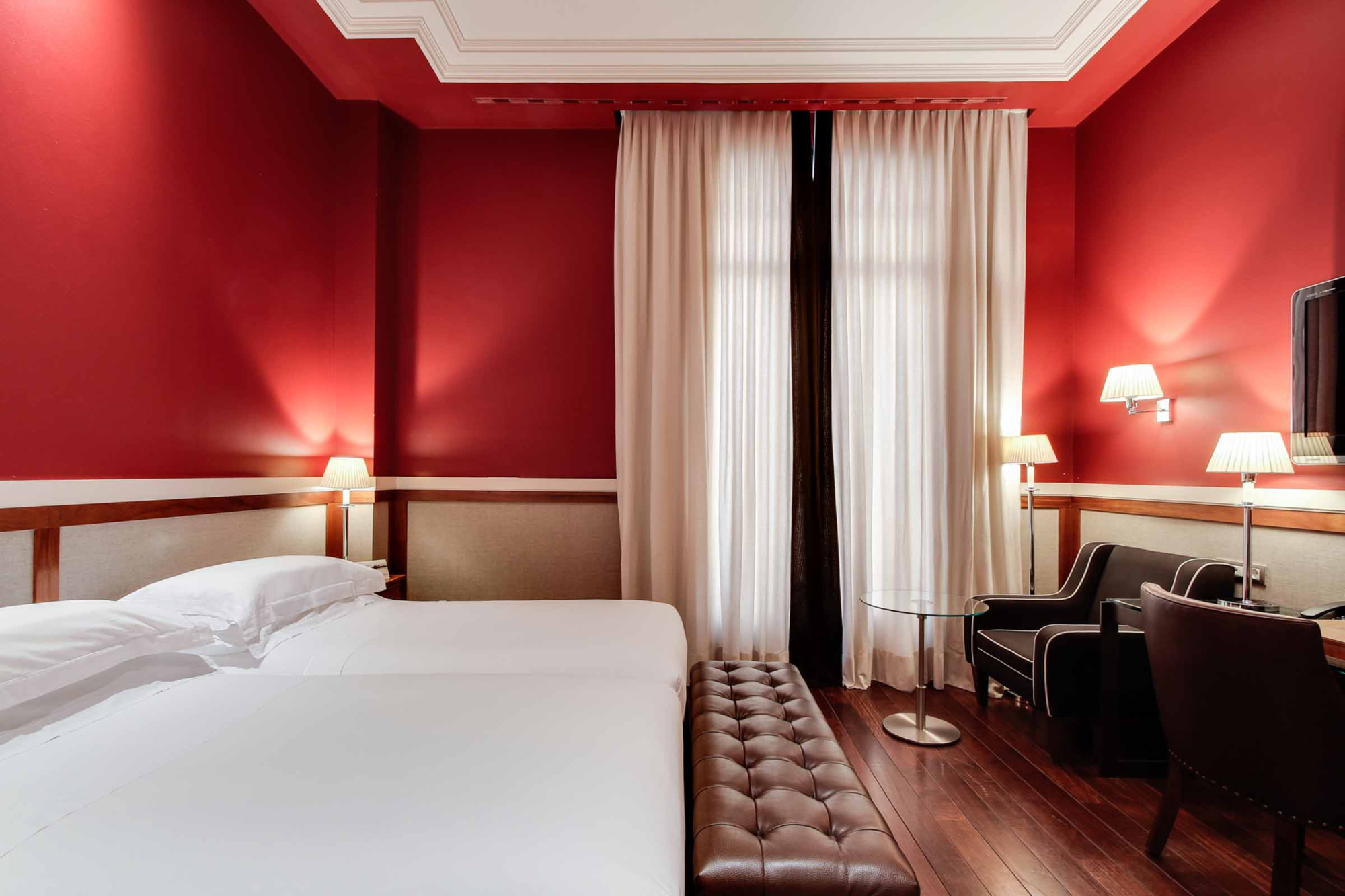 Se ve una cama doble, cortinas y una zona de estar con sillón y mesa auxiliar en la habitación.