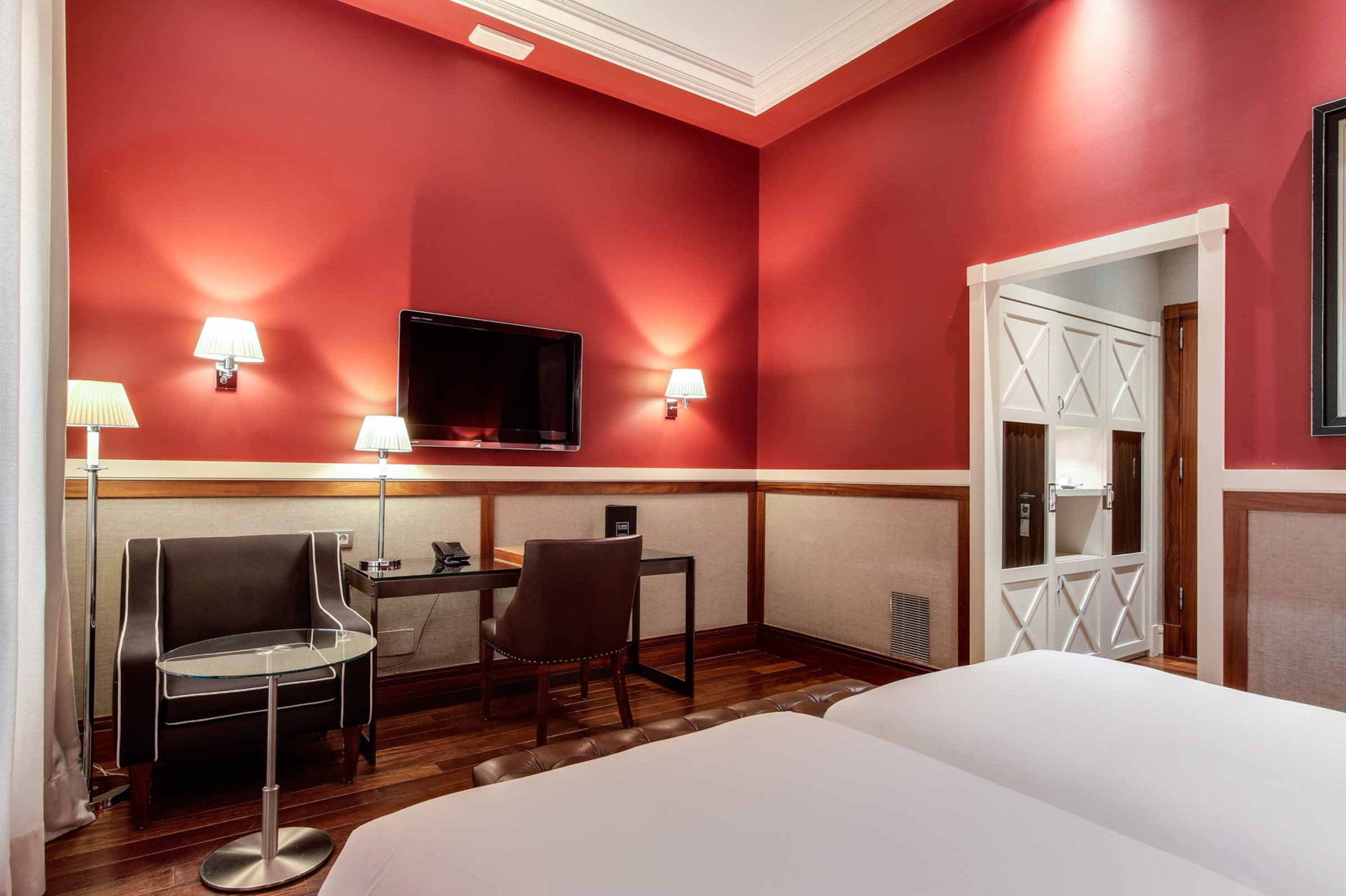 Se ve una cama, un televisor y una zona de estar con sillas y lámparas en una habitación con paredes rojas.