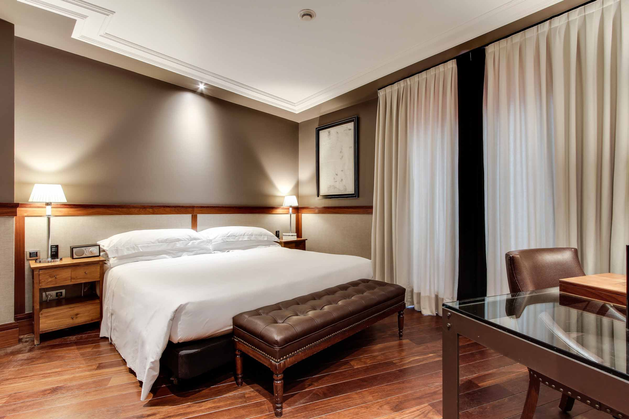 Se ve una habitación con una cama doble, mesitas de noche y cortinas junto a una zona de estar.