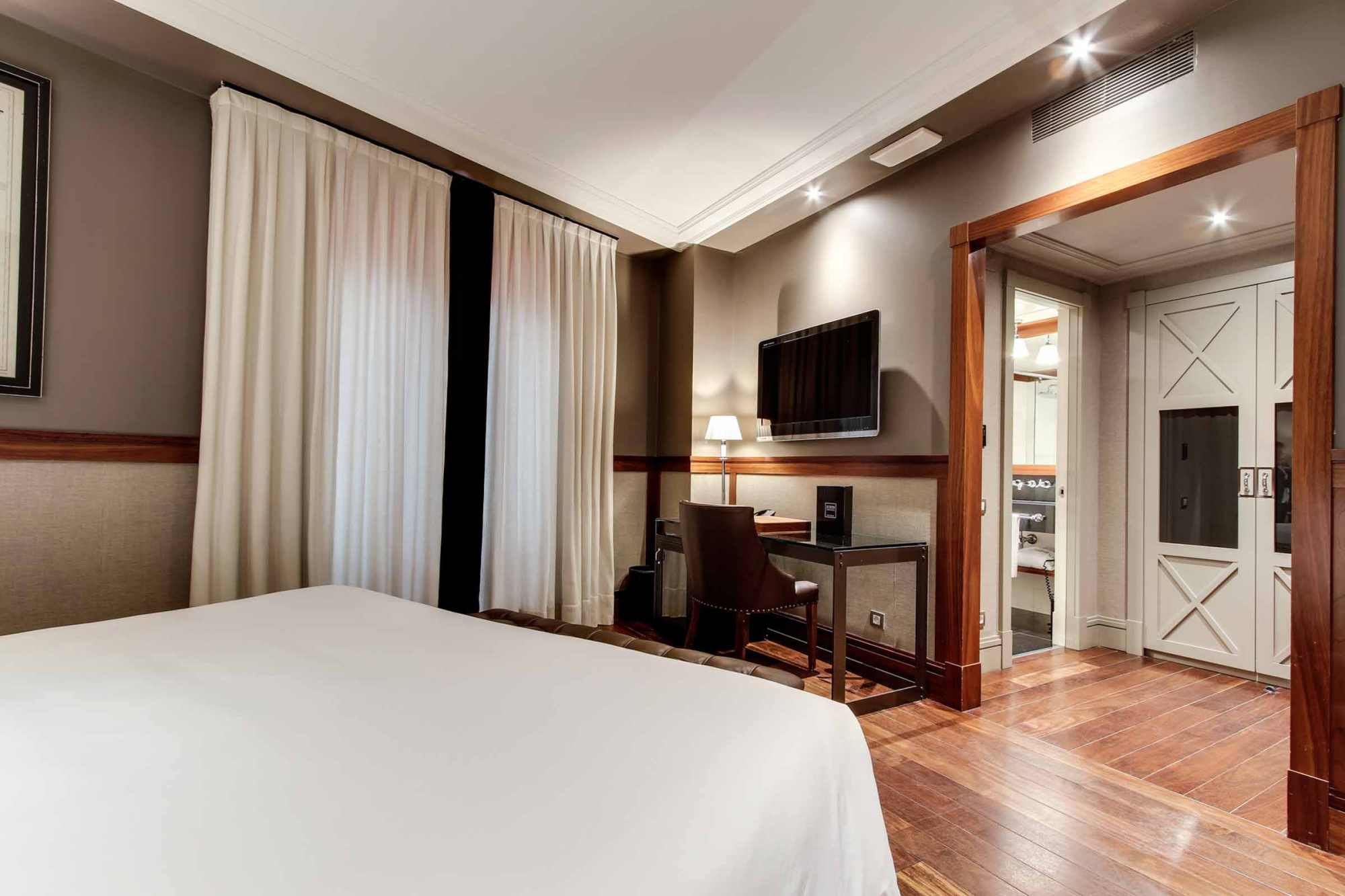 La habitación muestra una cama, cortinas, una televisión y un escritorio junto a una puerta interior.