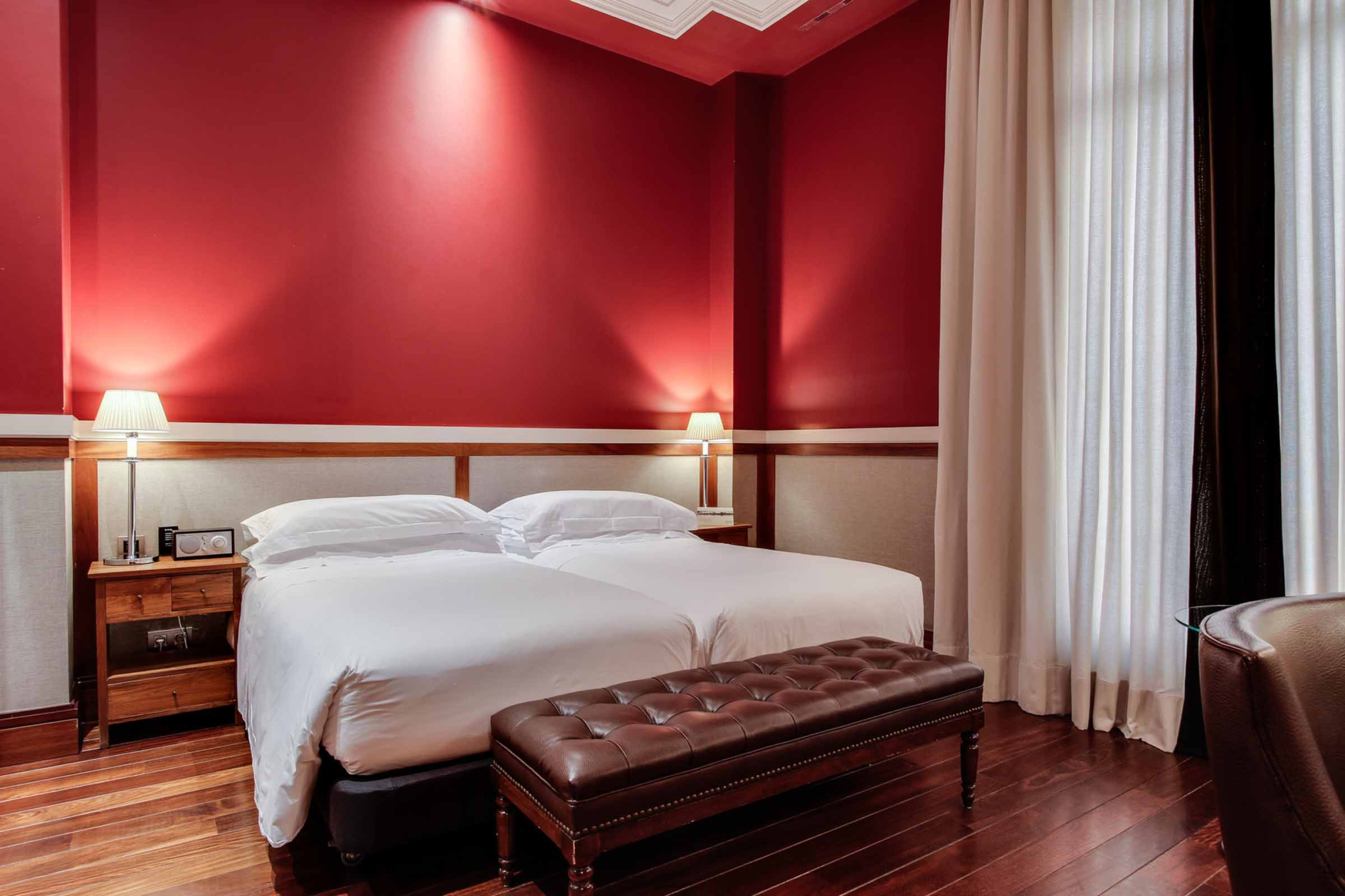 Se ve una cama doble con ropa blanca, mesitas y cortinas junto a una pared roja.