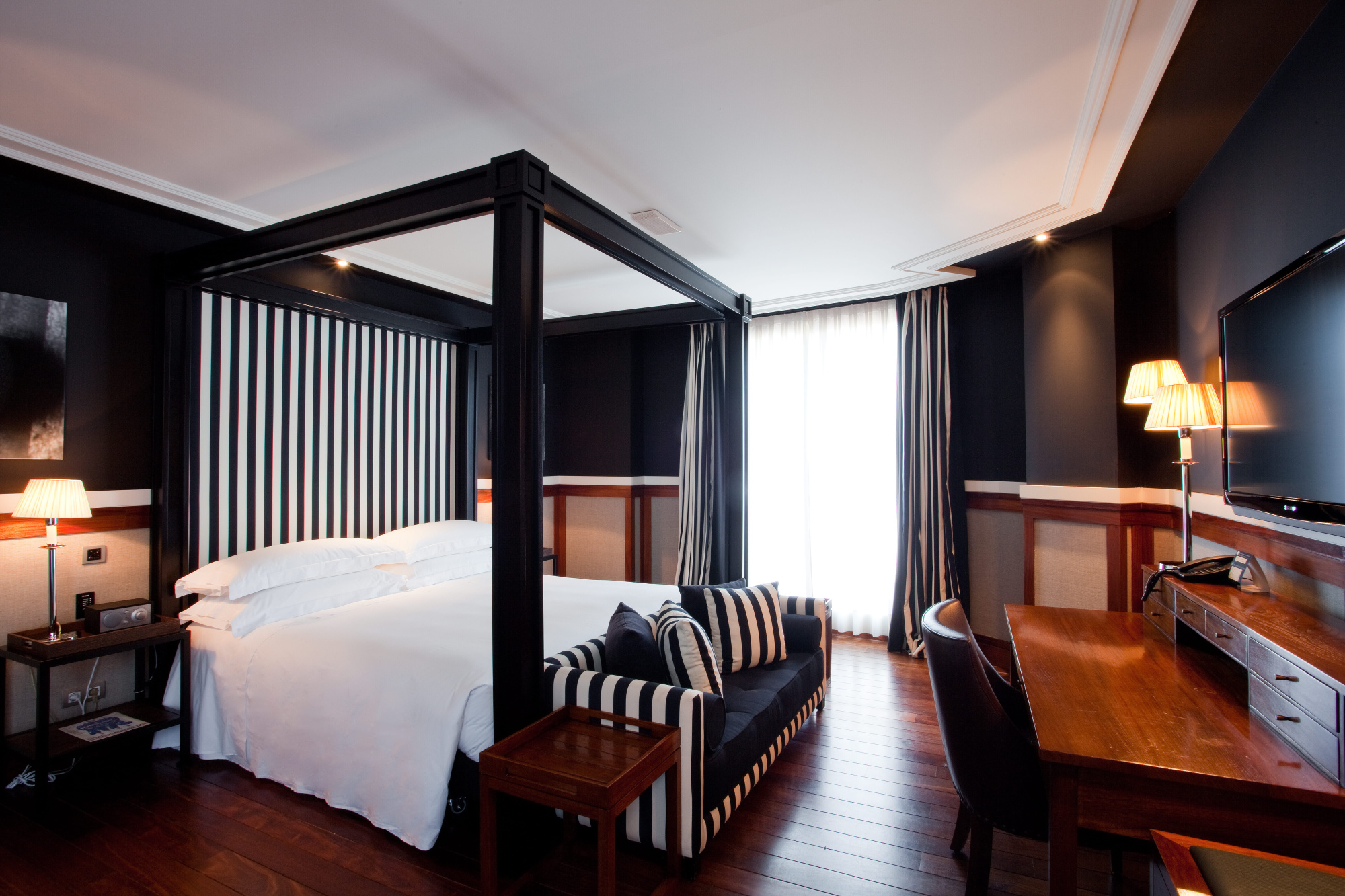 La imagen muestra una junior suite con cama, sillón con rayas, escritorio y lámparas de pie.