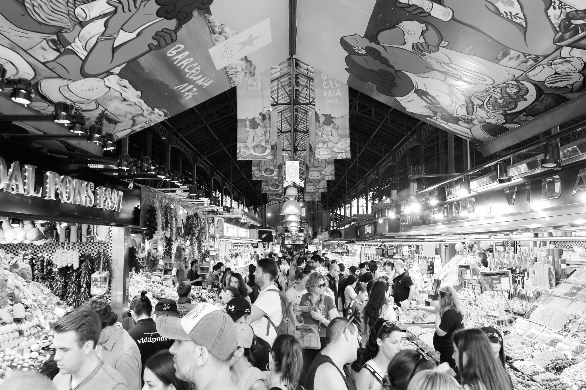 Se ve un mercado interior con muchos puestos y personas comprando bajo una estructura metálica.