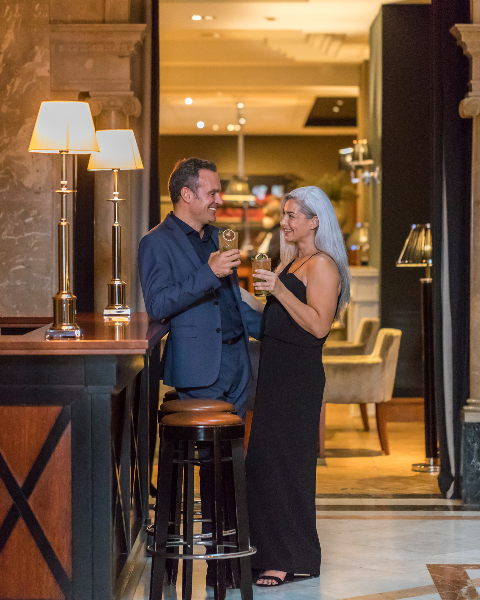 Una pareja brinda con copas en un vestíbulo de hotel con mesas y lámparas encendidas.