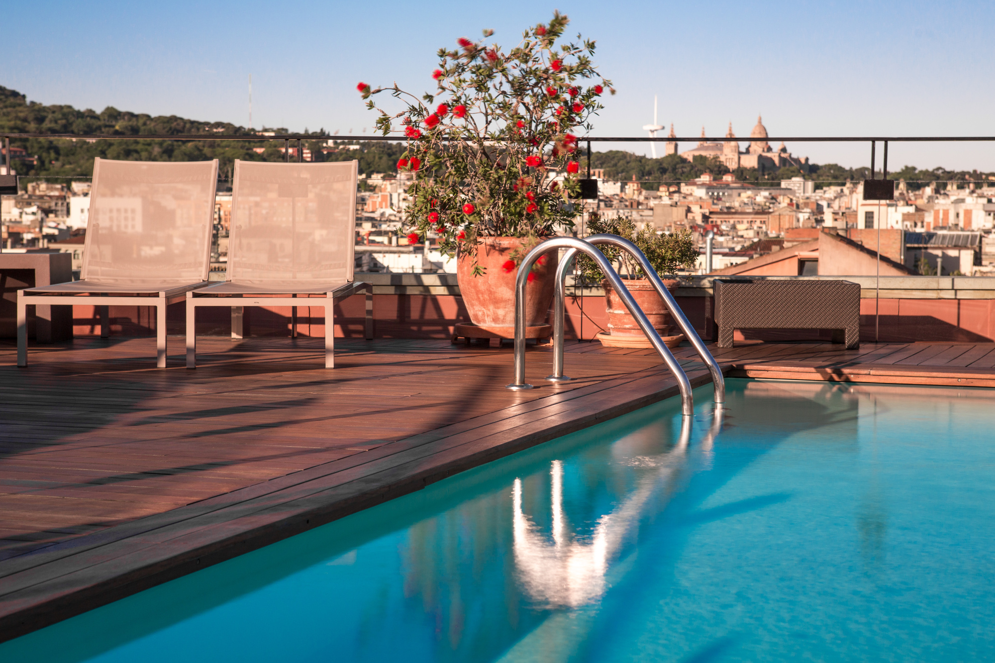 Se ve una piscina al aire libre con tumbonas y una barandilla de acero, con una ciudad al fondo.