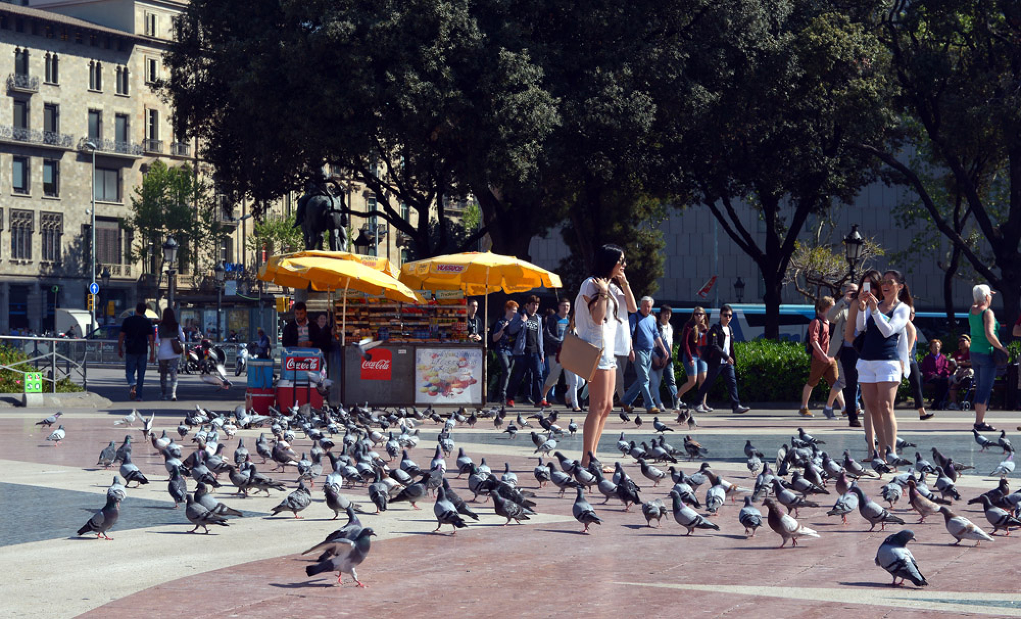 Una plaza urbana con palomas en el suelo y personas caminando y sentadas cerca de puestos.
