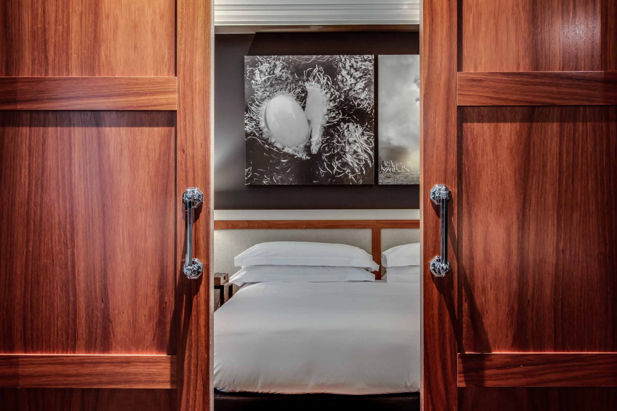 Se ve una puerta de madera abierta que da paso a una cama en un dormitorio con ropa de cama blanca.