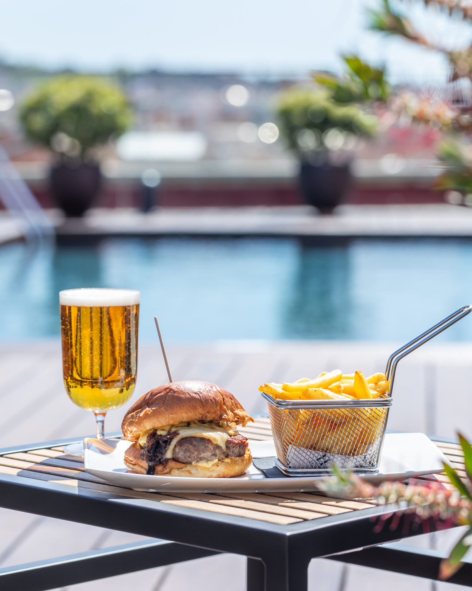 Sirve una mesa exterior con una hamburguesa, patatas, una jarra de cerveza y vasos sobre una terraza.
