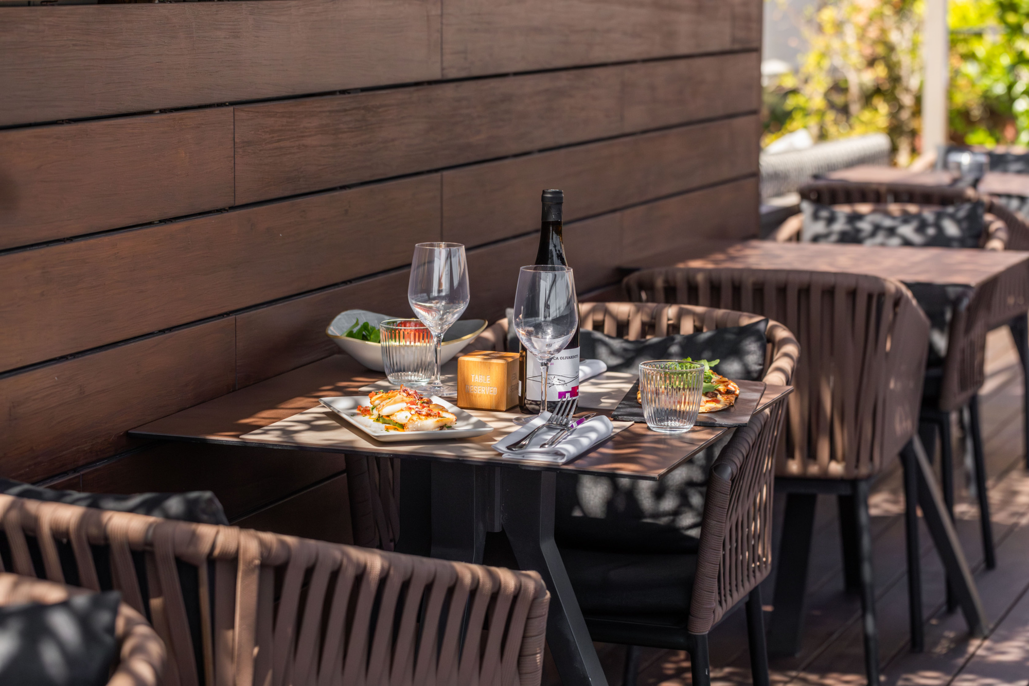 Se ve una mesa con copas y platos sobre un mantel, con sillas alrededor y un muro de fondo.