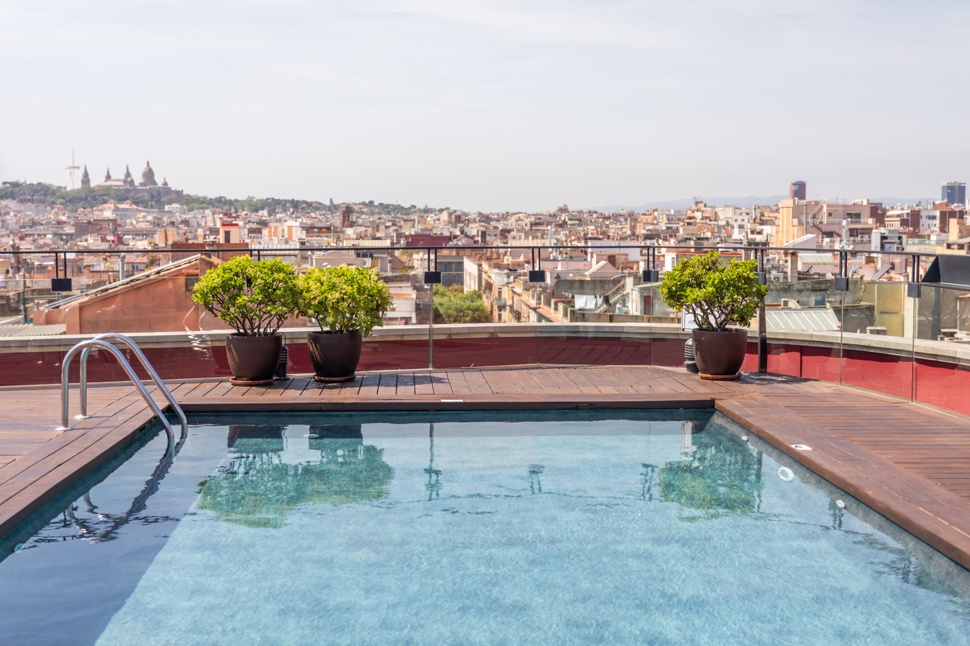 Se ve una piscina en una terraza con barandillas y macetas, con la ciudad al fondo.