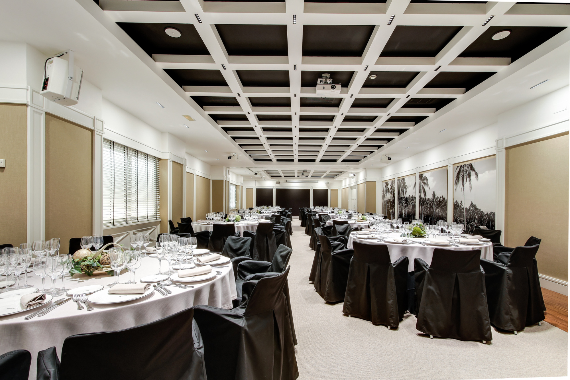 Presenta una sala de banquetes con mesas cubiertas y sillas alrededor, con iluminación en el techo.