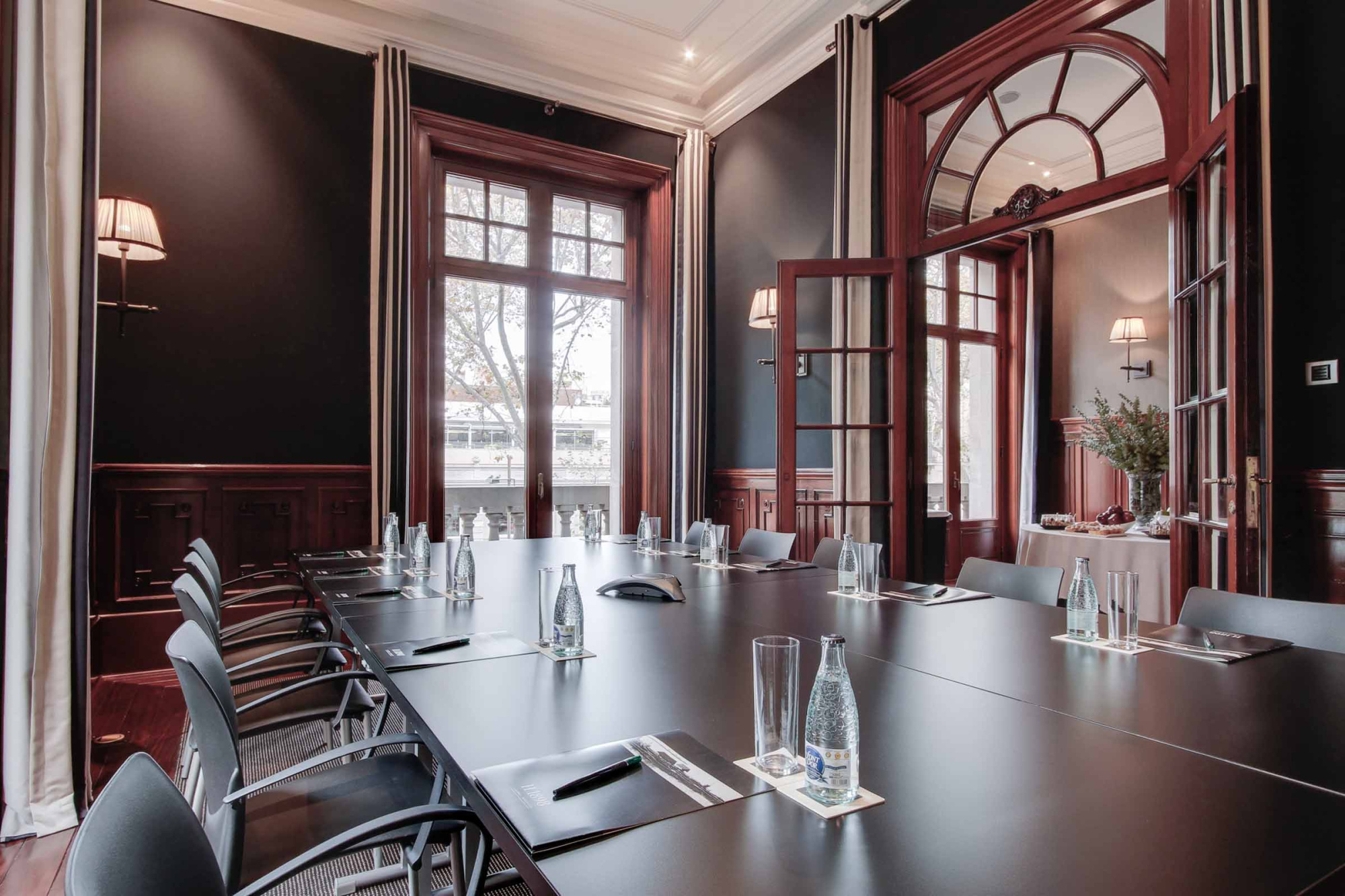La sala de reuniones muestra una mesa larga con sillas y elementos de mesa, junto a ventanas y lámparas.