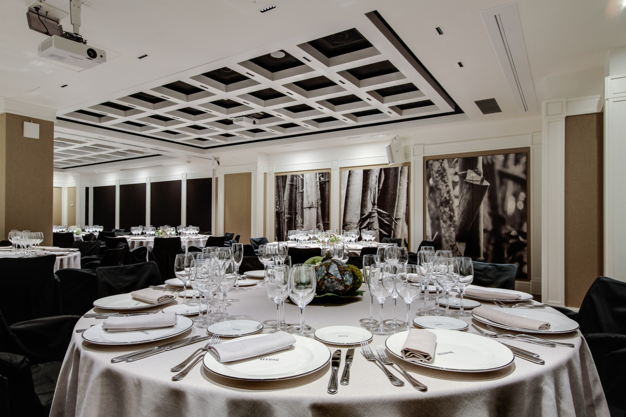 Presenta una mesa con vajilla y copas en un comedor con paredes decoradas y sillas alrededor.
