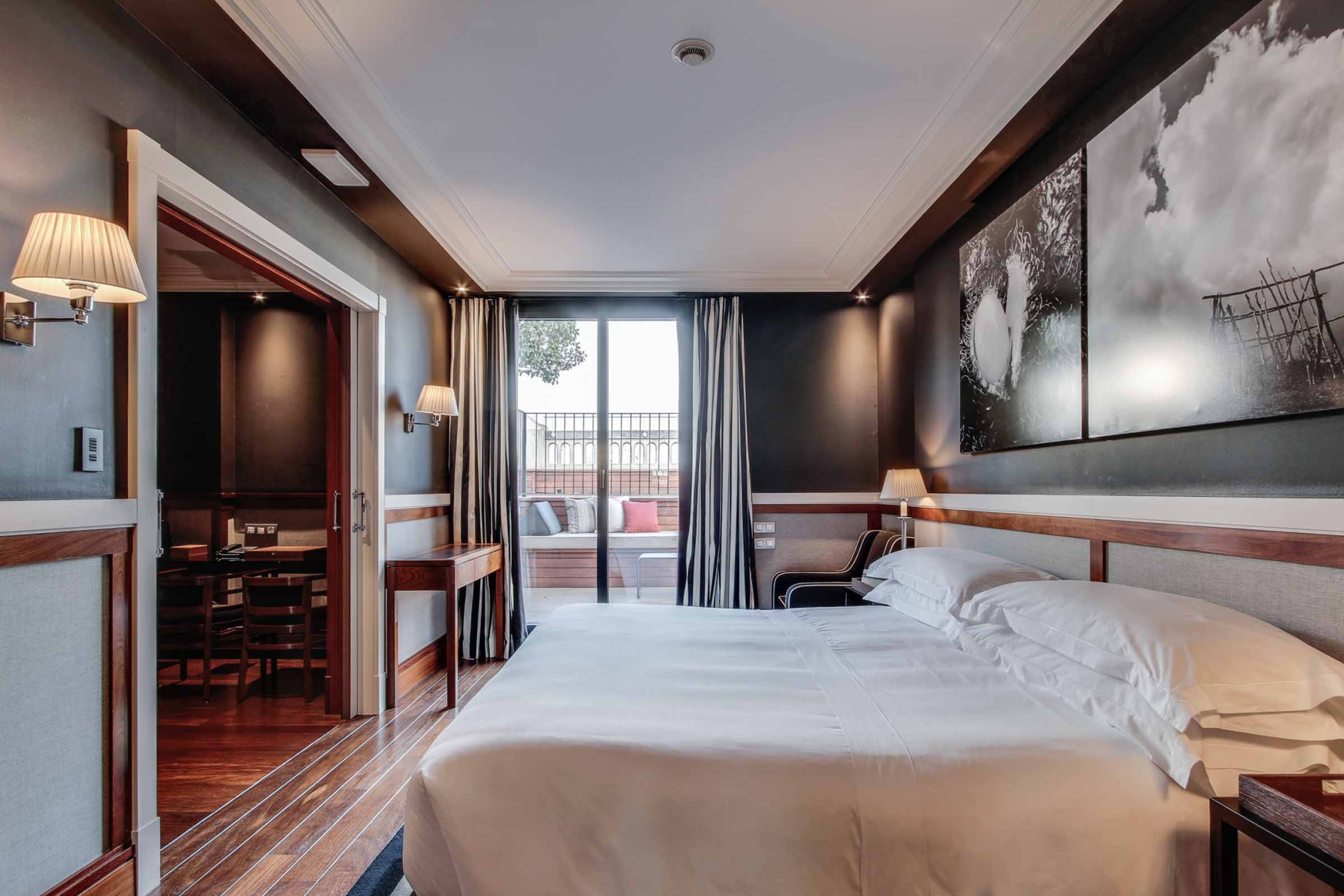 La imagen muestra una suite con cama doble, cortinas y una zona de estar con mesa y sillas.