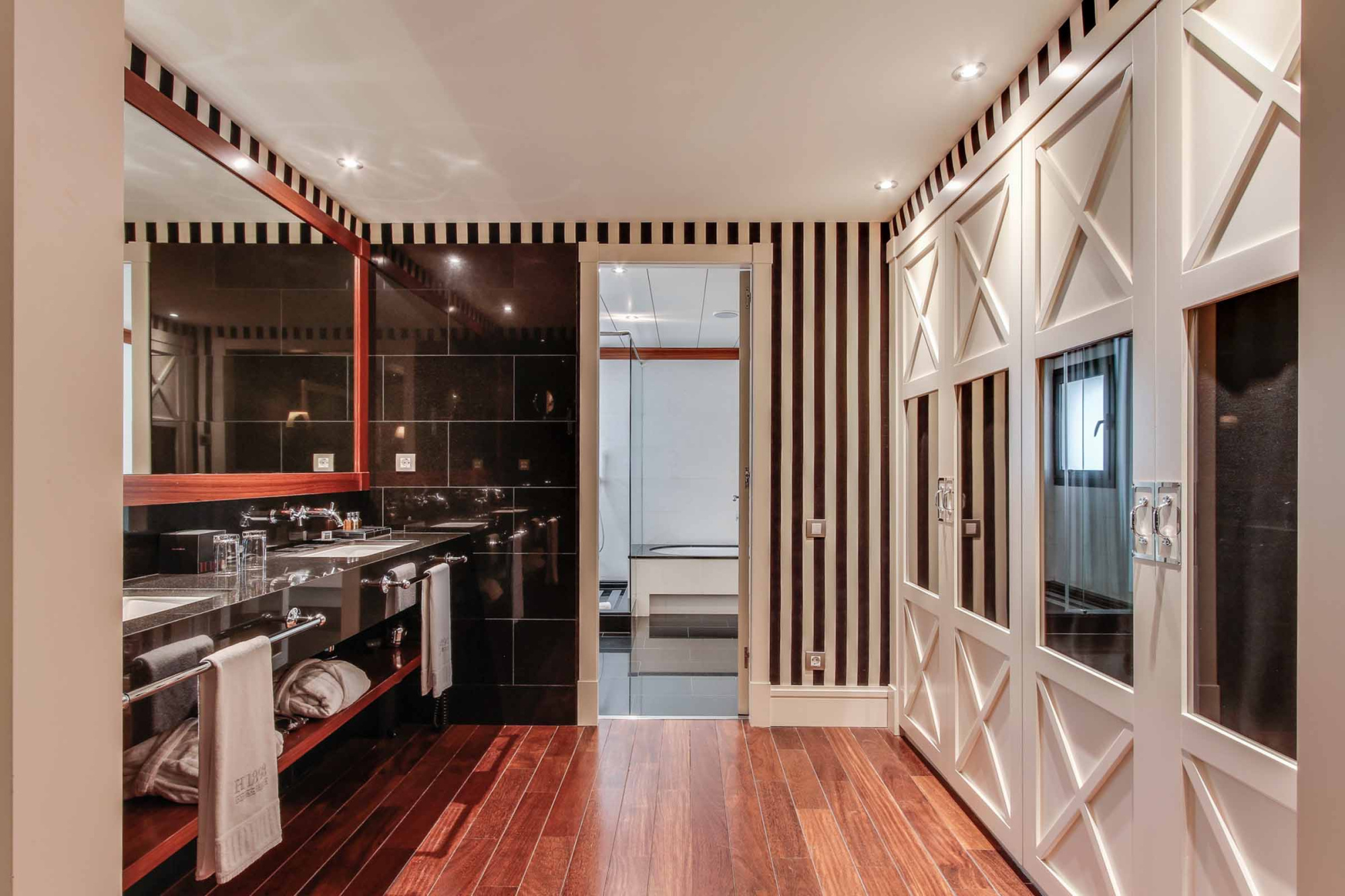 Se ve una suite con suelo de madera, paredes con paneles y una zona de tocador con lavabo y espejo.