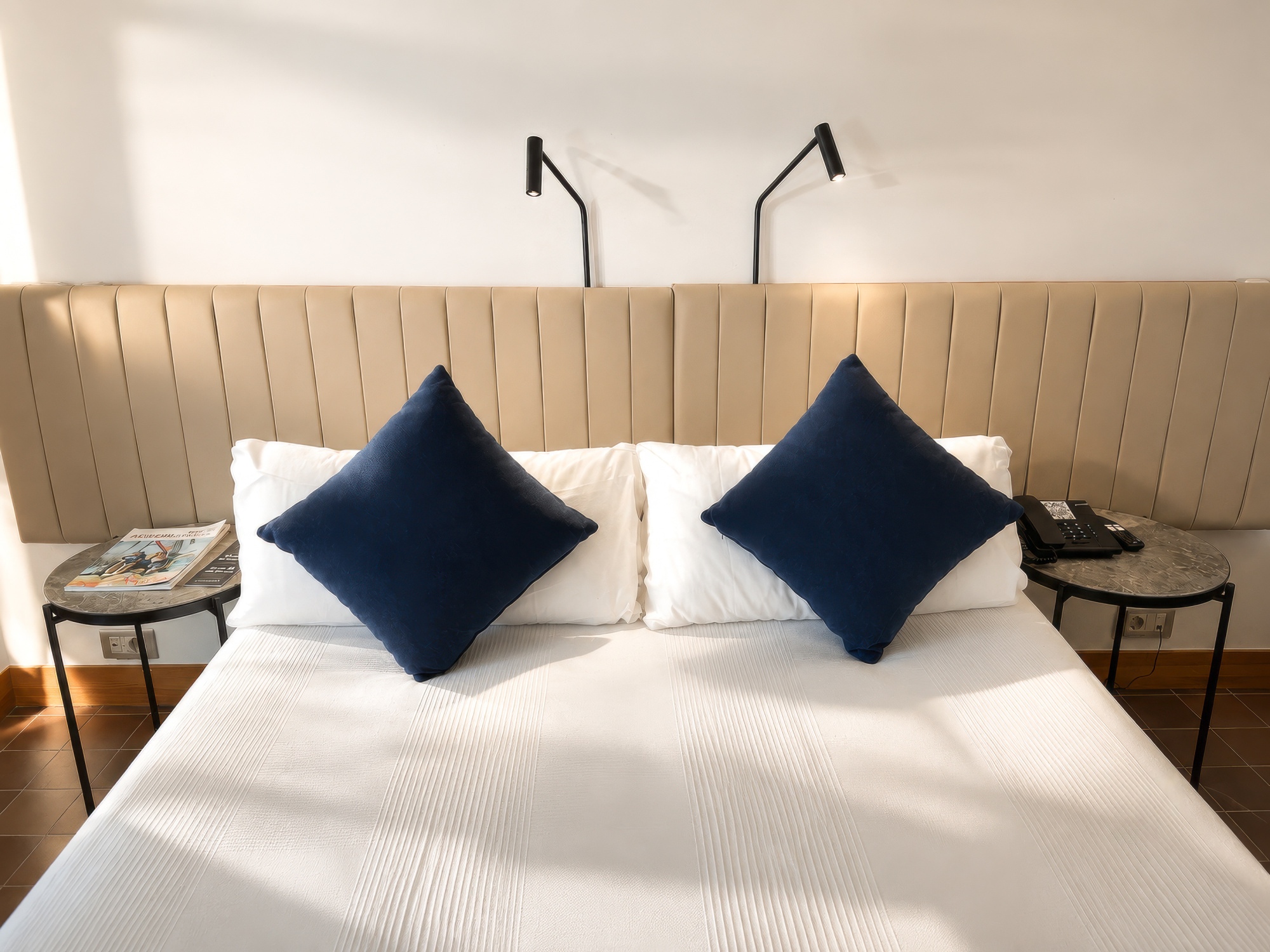 Se ve una cama doble con ropa de cama blanca y dos cojines azules sobre un cabecero tapizado.