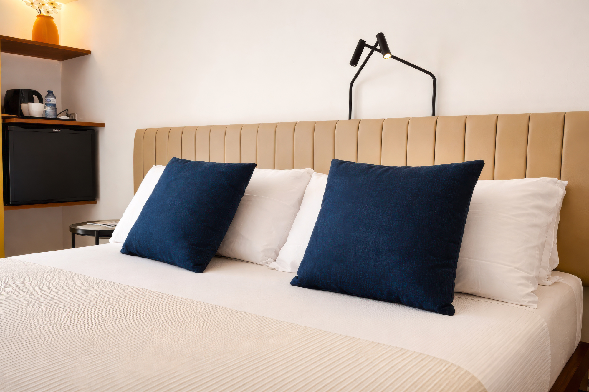 Se ve una cama doble con ropa de cama clara y dos almohadas azules sobre un cabecero de tela claro.